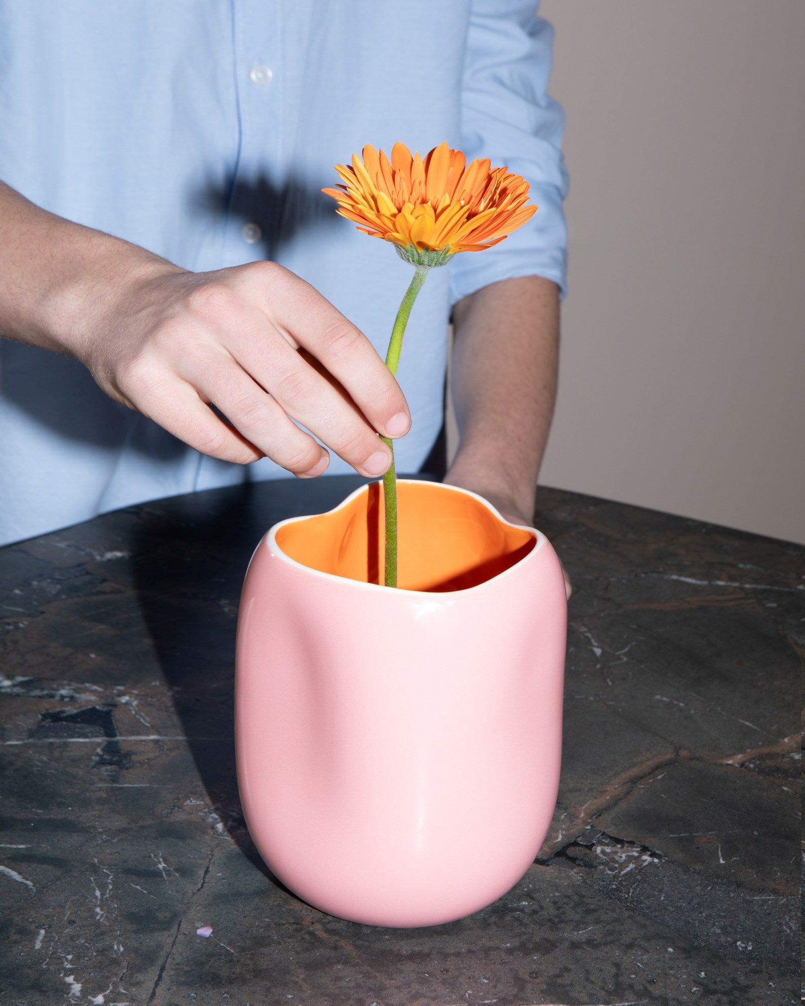 Hand platziert orangefarbene Blume in rosafarbener Vase auf Marmortisch.