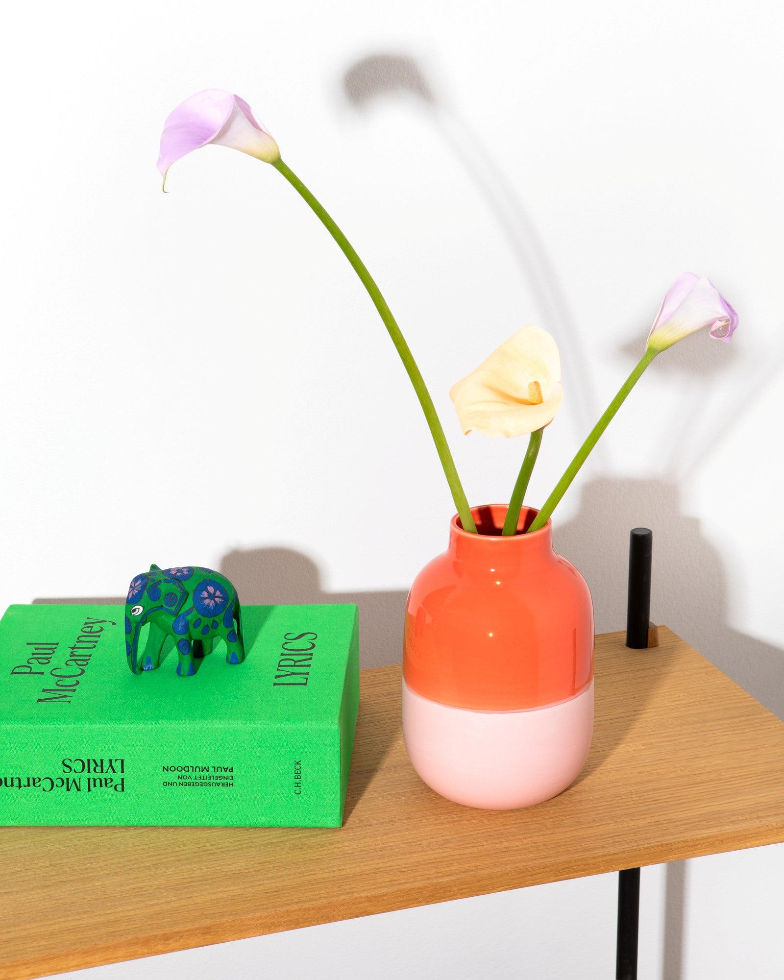 Eine bunte Vase mit Calla-Lilien steht auf einem Tisch neben einem grünen Buch mit einer kleinen Elefantenfigur darauf.