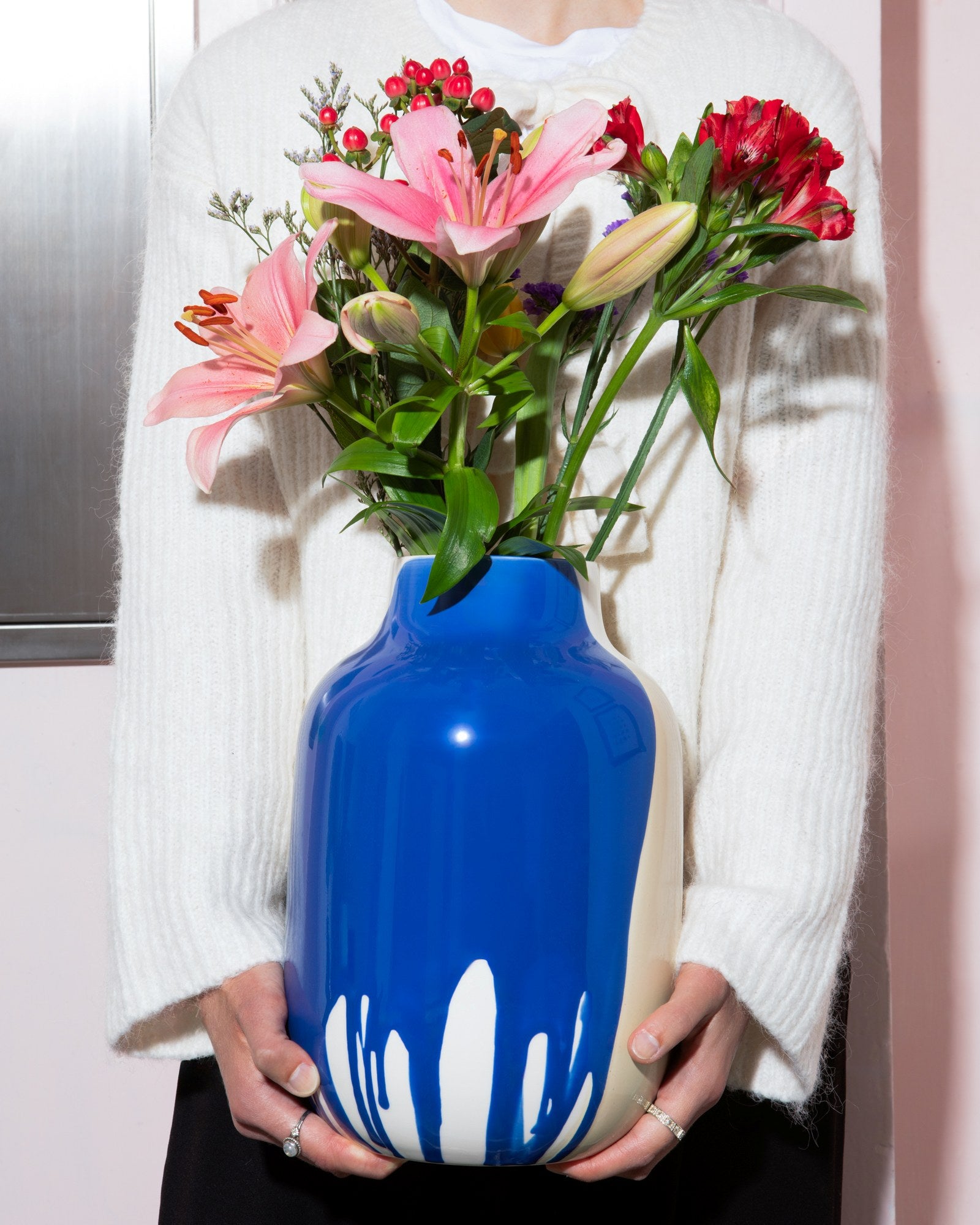 Person hält eine blau-weiße Vase mit rosa und roten Blumen vor einem weißen Pullover.