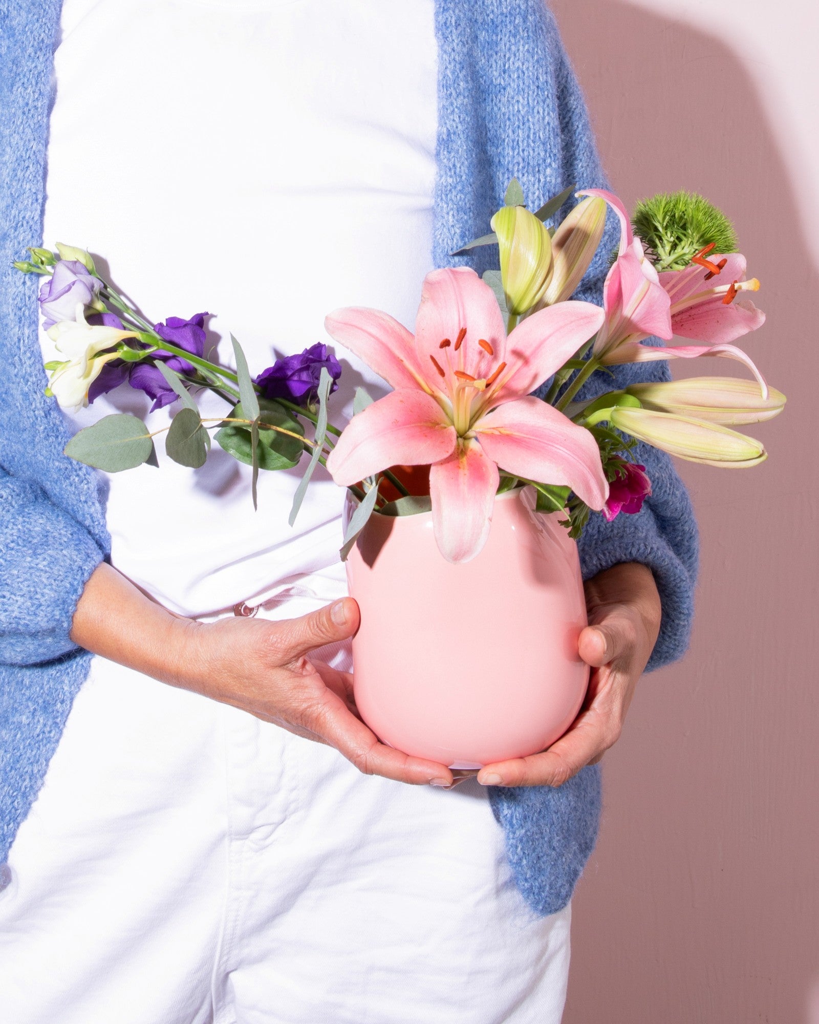 Eine Person hält eine rosa Vase mit bunten Blumen, trägt weiße Kleidung und eine blaue Strickjacke.