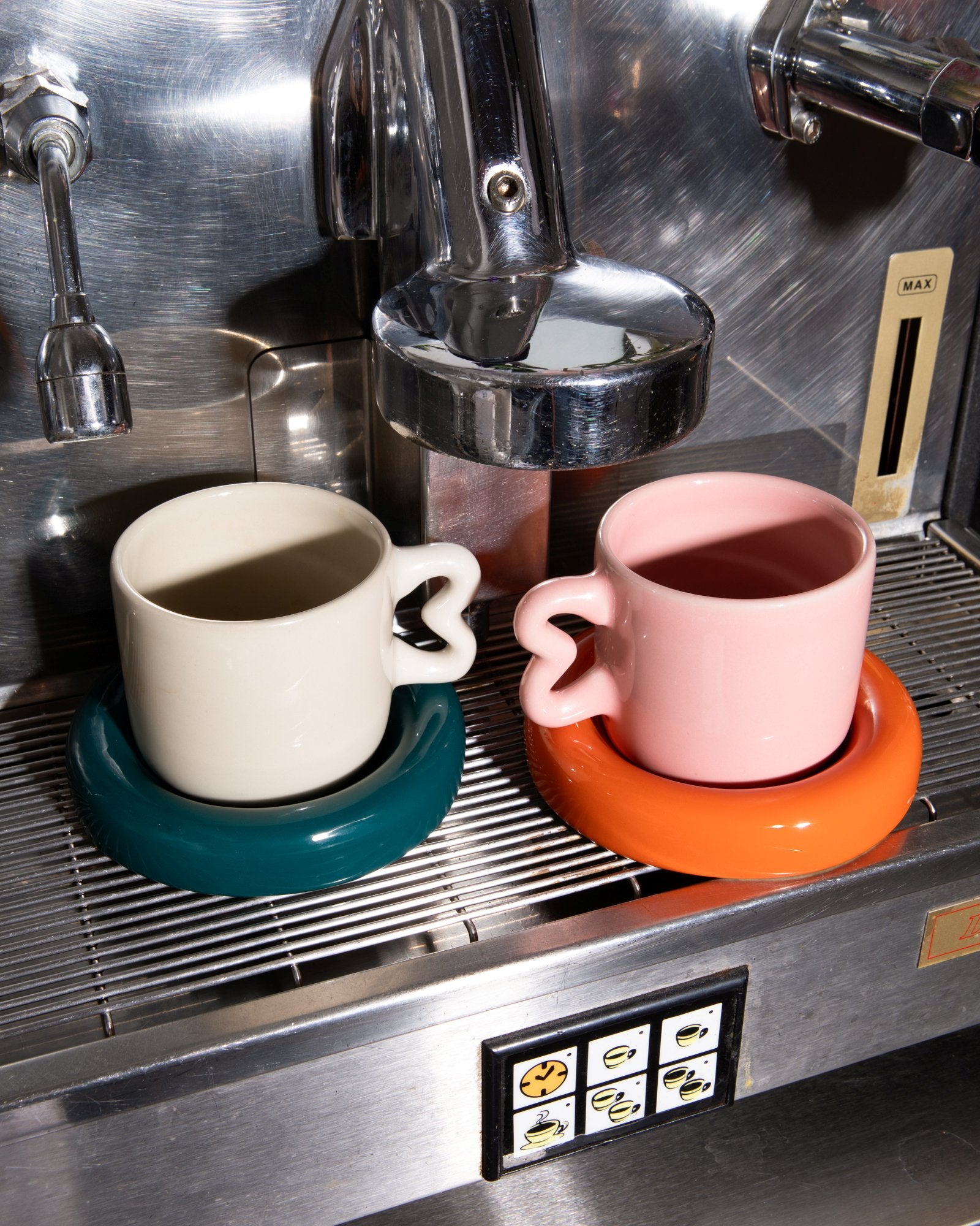 Zwei bunte Tassen auf einer Espressomaschine, eine beige mit grünem Untersatz und eine rosa mit orangem Untersatz.