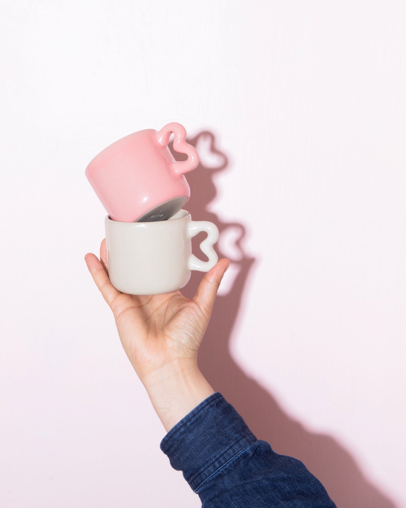 Eine Hand hält zwei übereinander gestapelte Tassen mit herzförmigen Henkeln vor einem rosa Hintergrund.