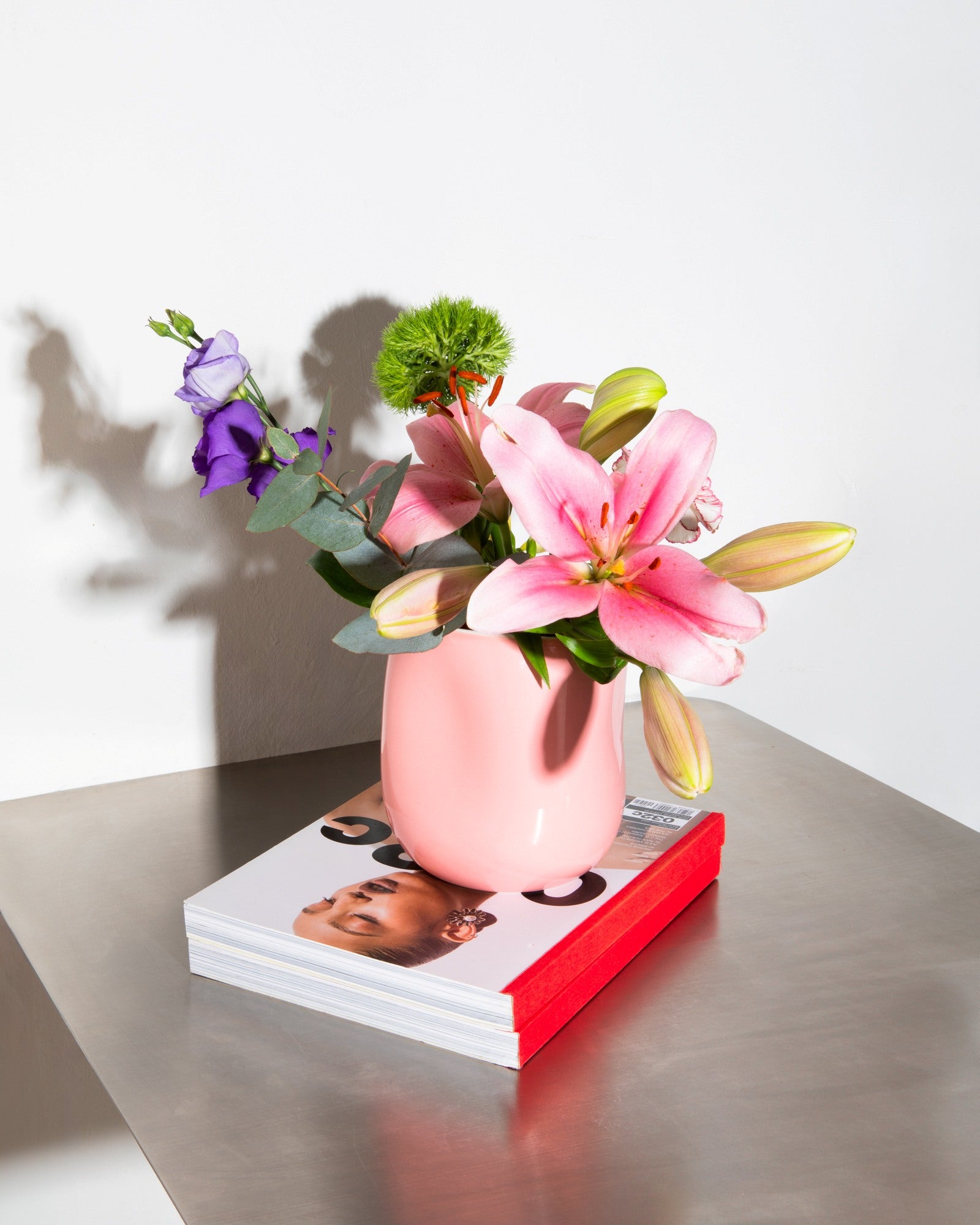Rosa Vase mit Blumenarrangement auf einem Tisch; darunter liegt ein Magazin mit rotem Umschlag.