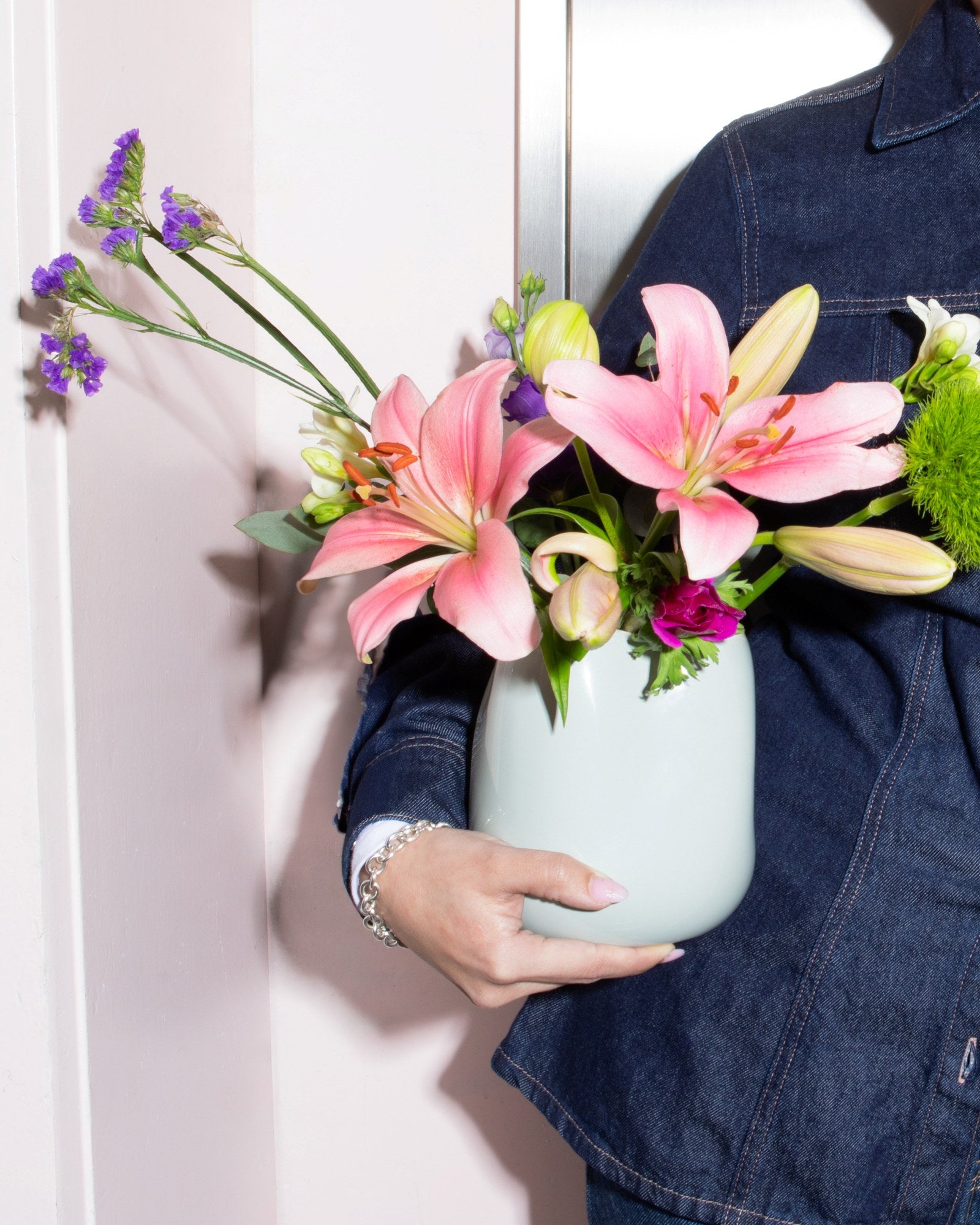 Person in Jeansjacke hält eine Vase mit rosa Blumen, darunter Lilien, und violetten Blüten vor pastellfarbenem Hintergrund.