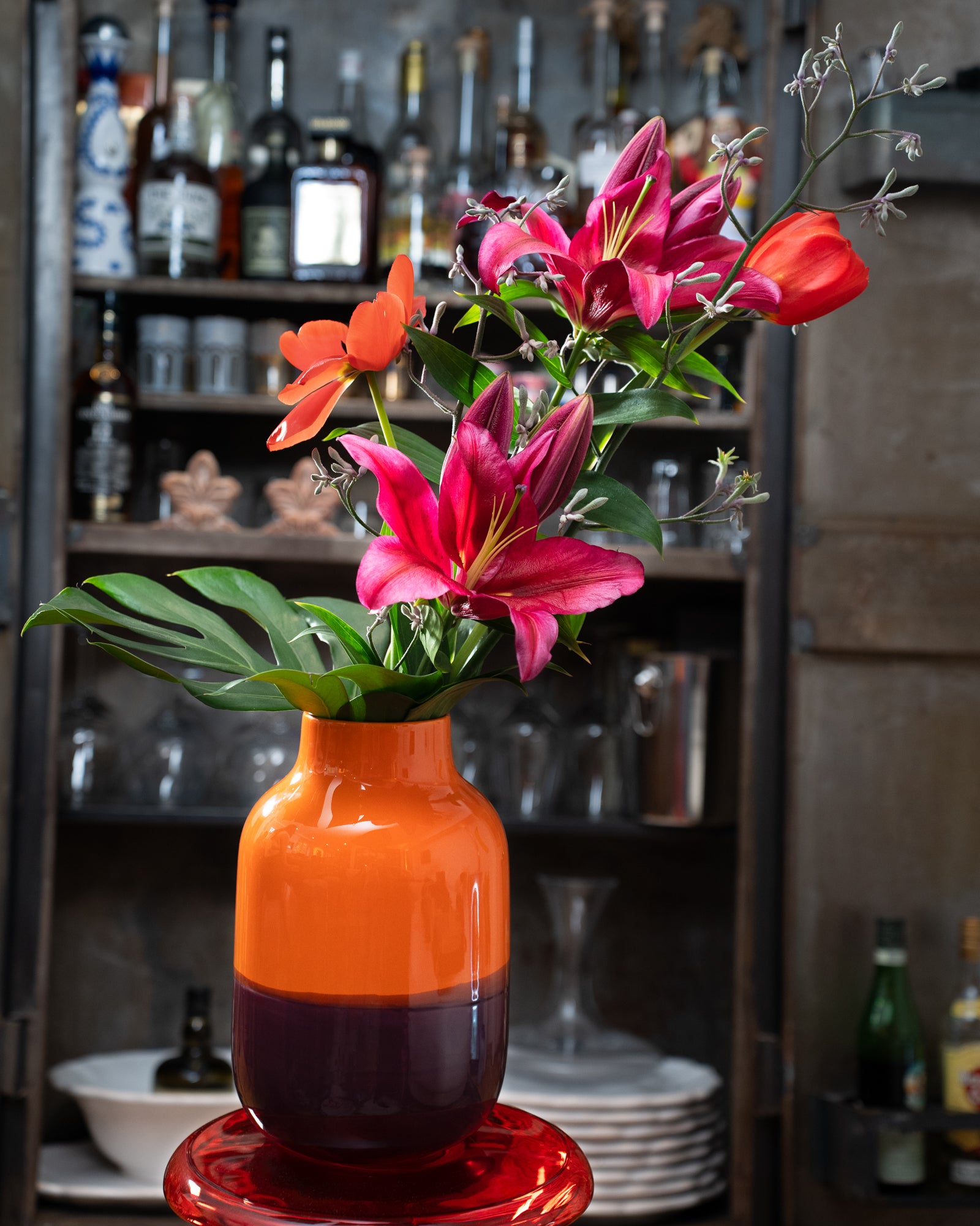 Ein orange-violette Vase mit pinken und roten Lilien steht vor einem Regal mit Flaschen und Gläsern.