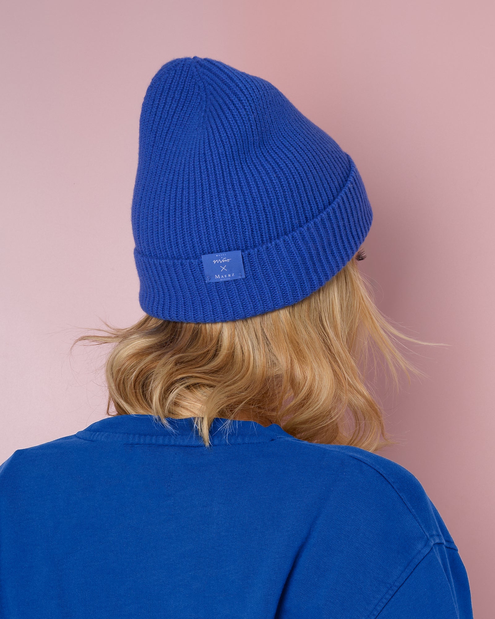 Person von hinten in einem blauen Pullover und blauer Mütze vor einem rosa Hintergrund.