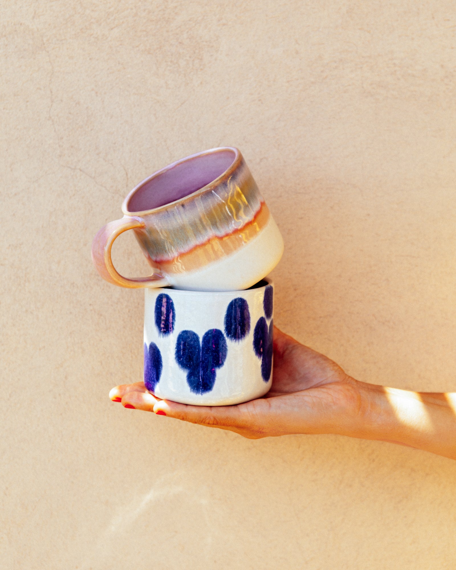 Zwei bunte Tassen werden von einer Hand vor einer beige Hintergrundwand gehalten.