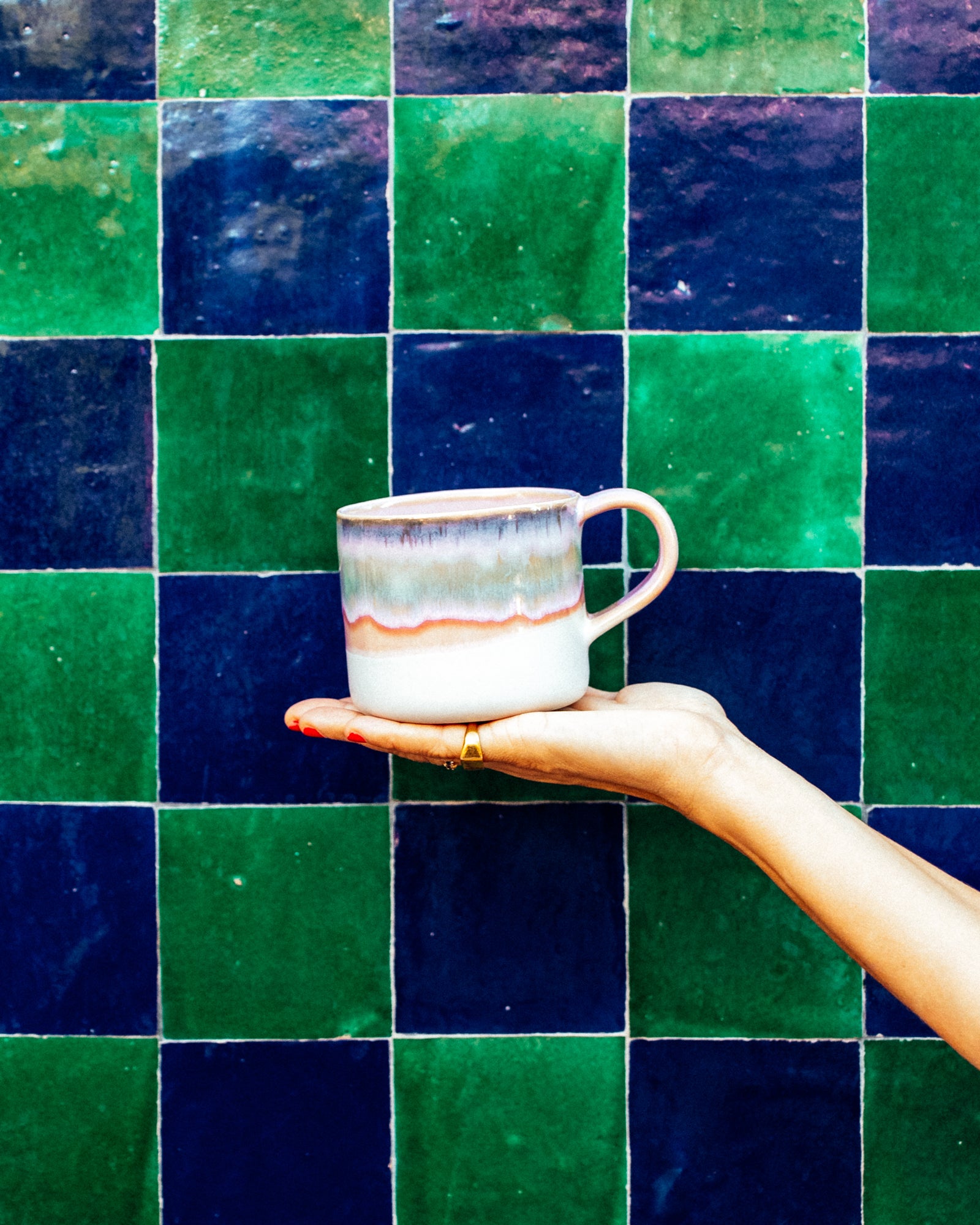 Eine Hand hält eine bunte Tasse vor einer Wand mit grünen und blauen Kacheln.
