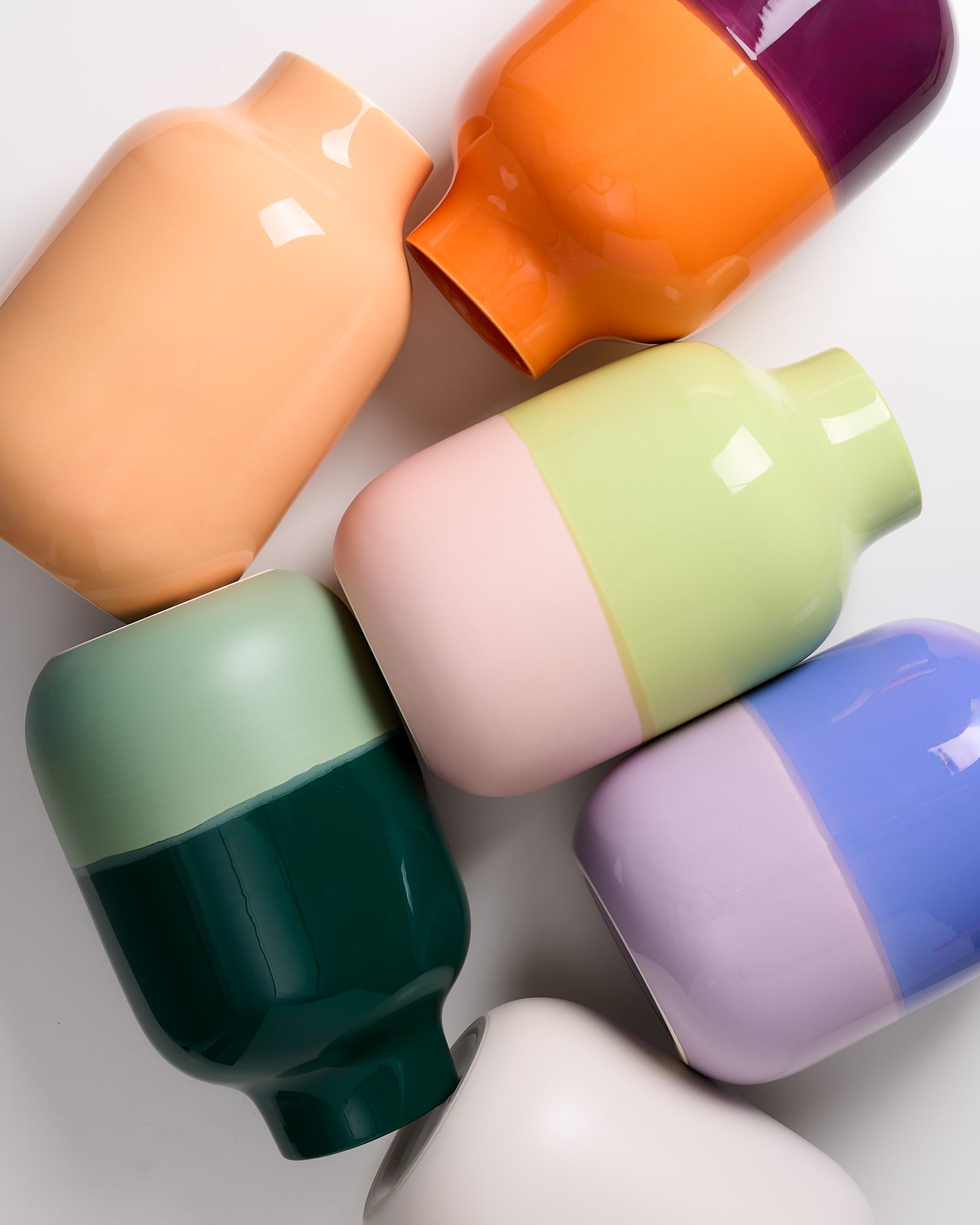 Sechs bunte, glänzende Vasen in verschiedenen Farben liegen nebeneinander auf einem weißen Hintergrund.