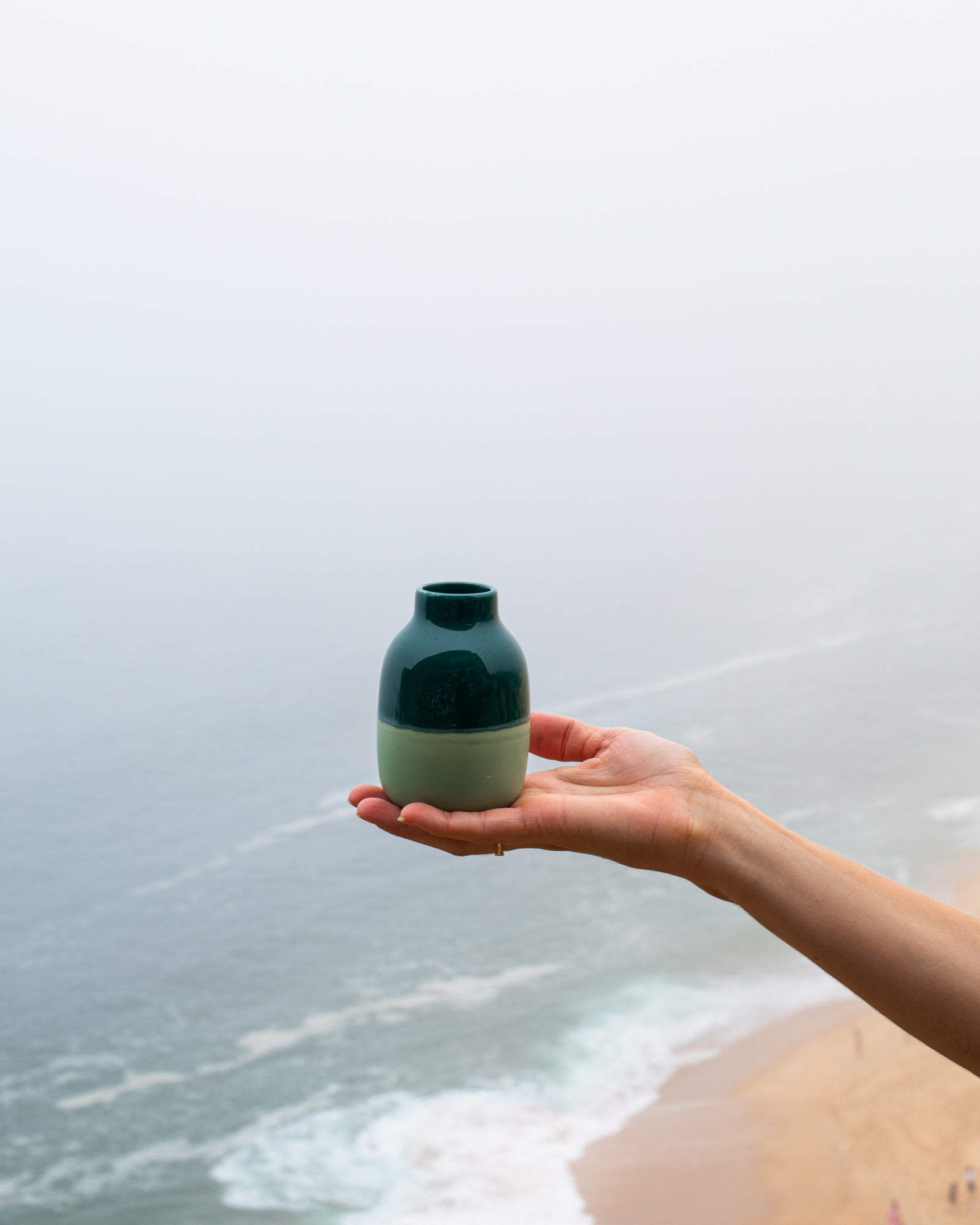 Eine Hand hält eine grünliche Vase vor einem verschwommenen Meereshintergrund.