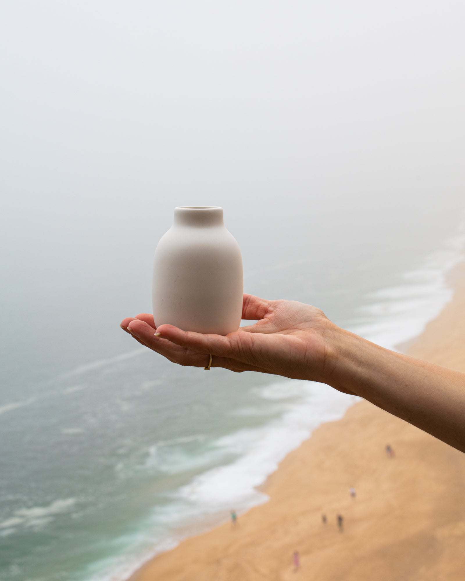 Eine Hand hält eine weiße Vase über einem nebligen Strand mit Wellen im Hintergrund.