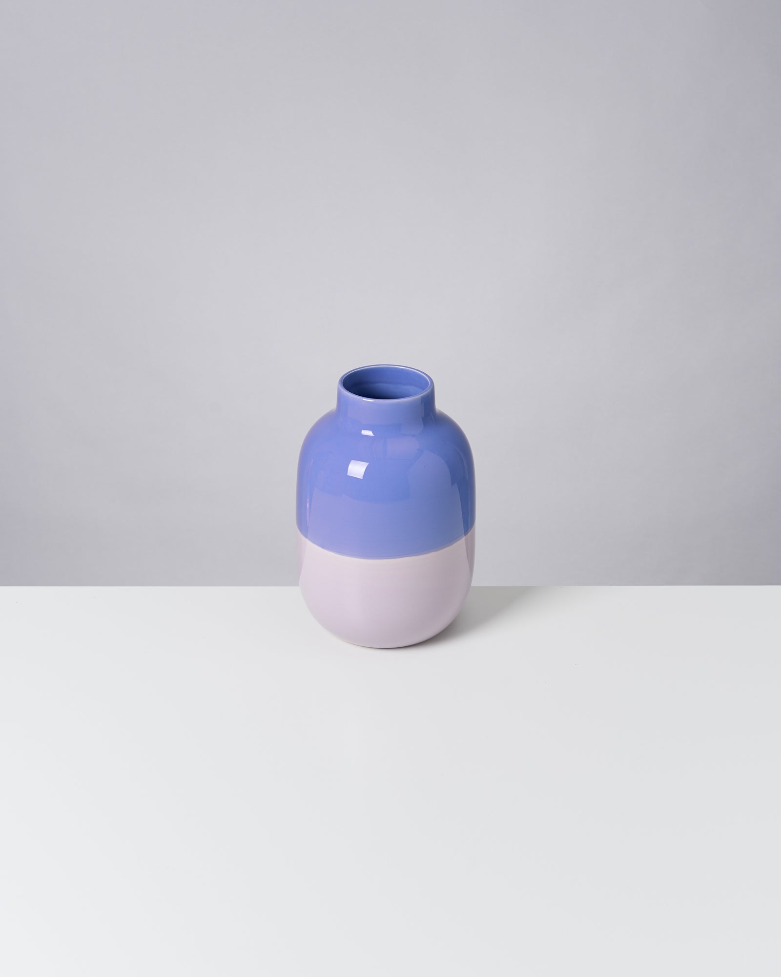 Eine zweifarbige, bauchige Vase in Blau und Hellrosa steht auf einem weißen Tisch vor grauem Hintergrund.