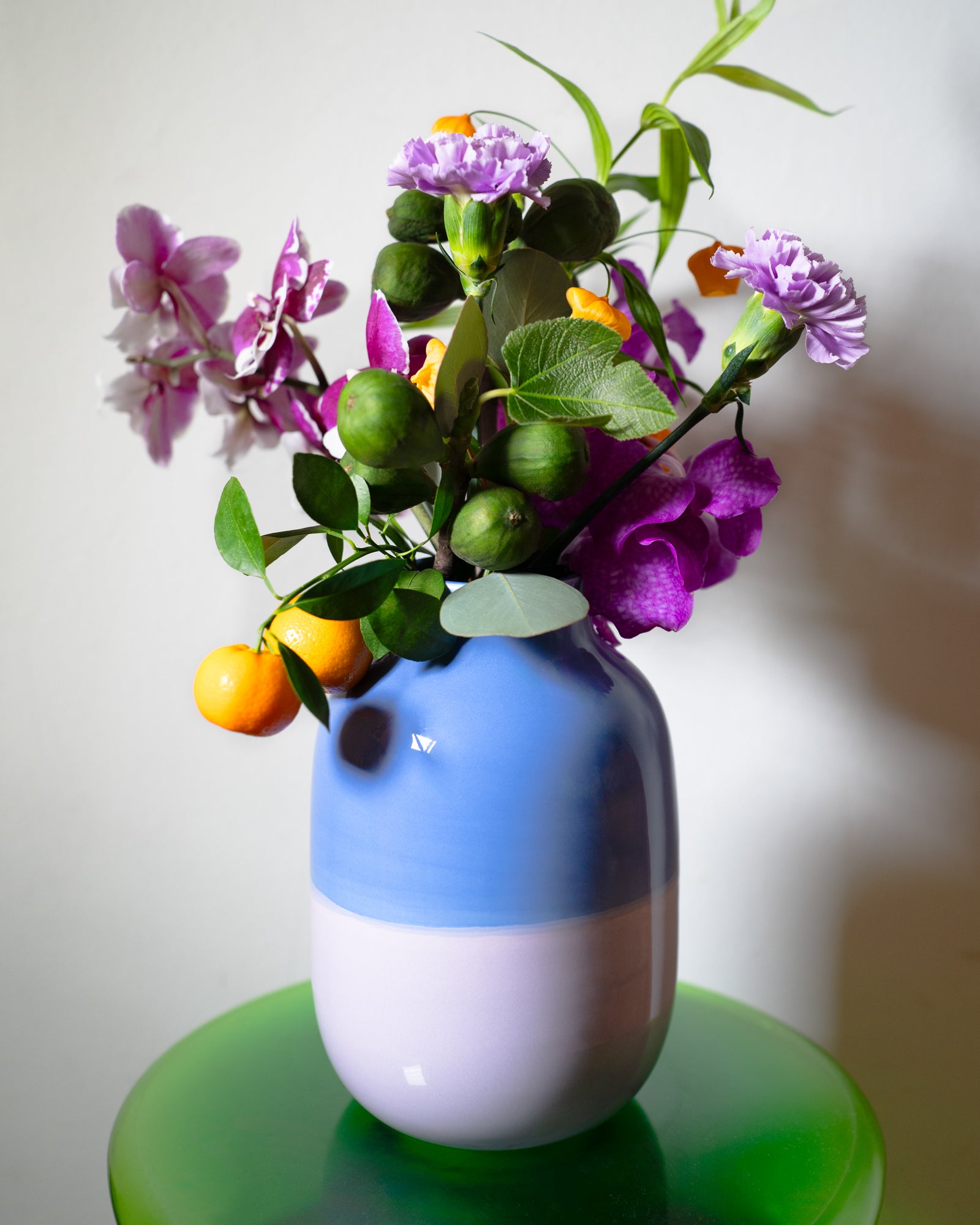 Bunter Blumenstrauß mit lila Blüten und Orangen in einer blauen Vase auf einem grünen Tisch.