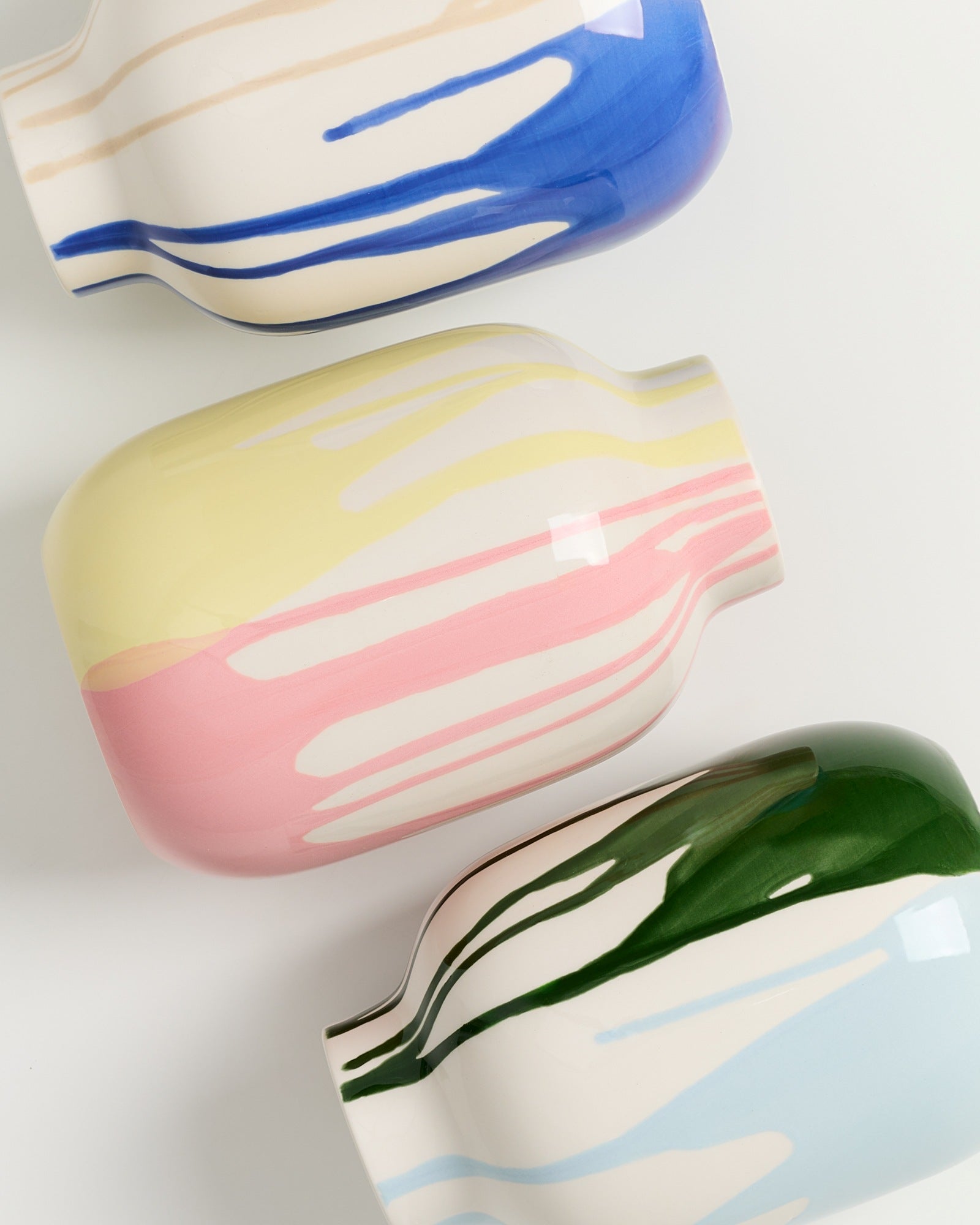 Drei bunte Vasen mit blauen, rosa und grünen Streifen liegen nebeneinander auf einer weißen Fläche.