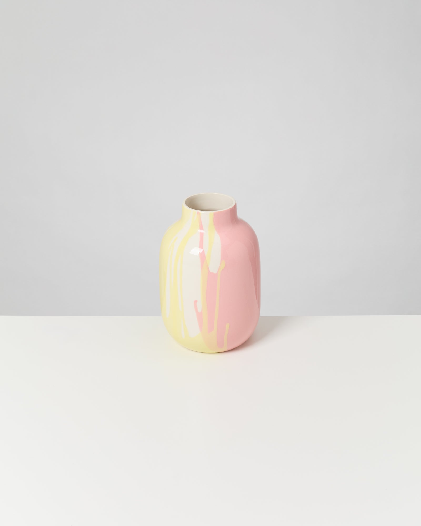 Eine Keramikvase mit rosa und gelbem Farbverlauf steht auf einem weißen Tisch vor grauem Hintergrund.