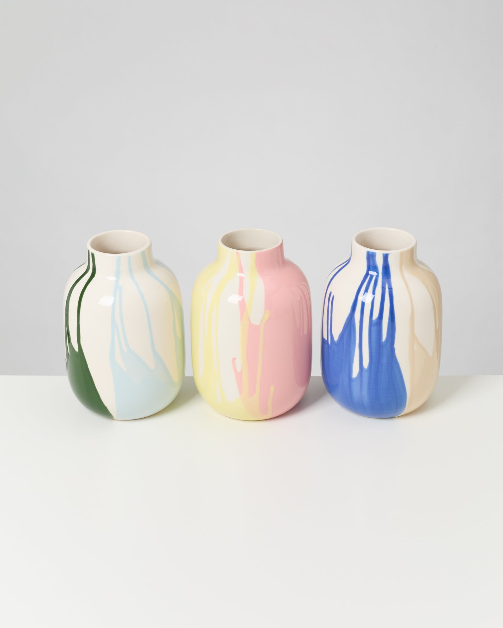 Drei Vasen mit bunten, tropfenden Mustern in Grün, Pink und Blau stehen nebeneinander auf einem weißen Tisch.