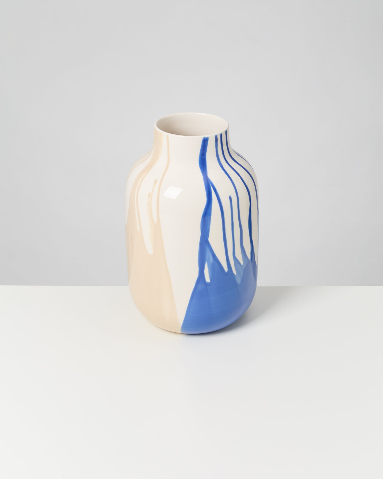Beigefarbene Vase mit blauen, tropfenden Streifen auf weißem Hintergrund.