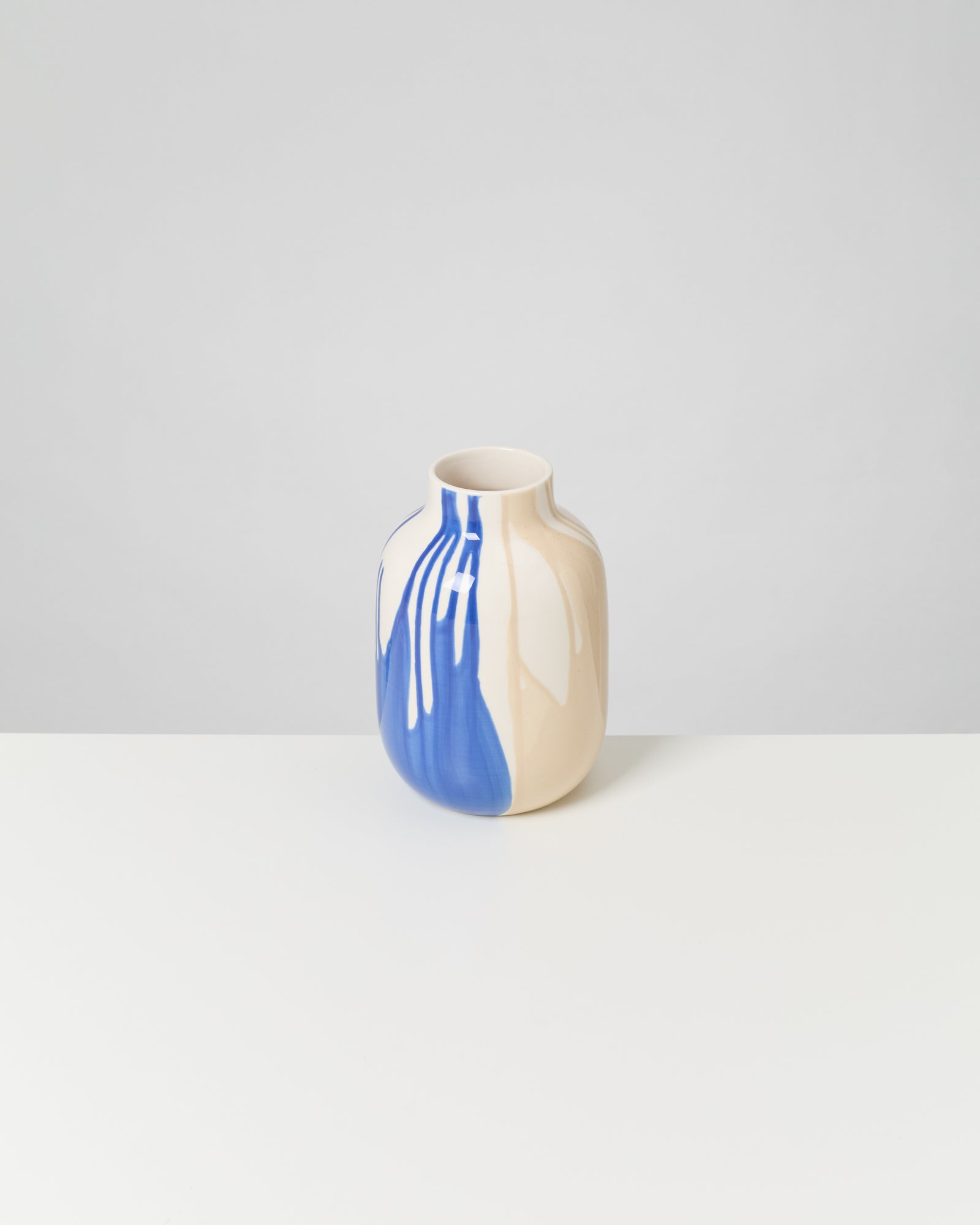 Beige Vase mit blauem, abstraktem Muster auf weißem Tisch vor grauem Hintergrund.