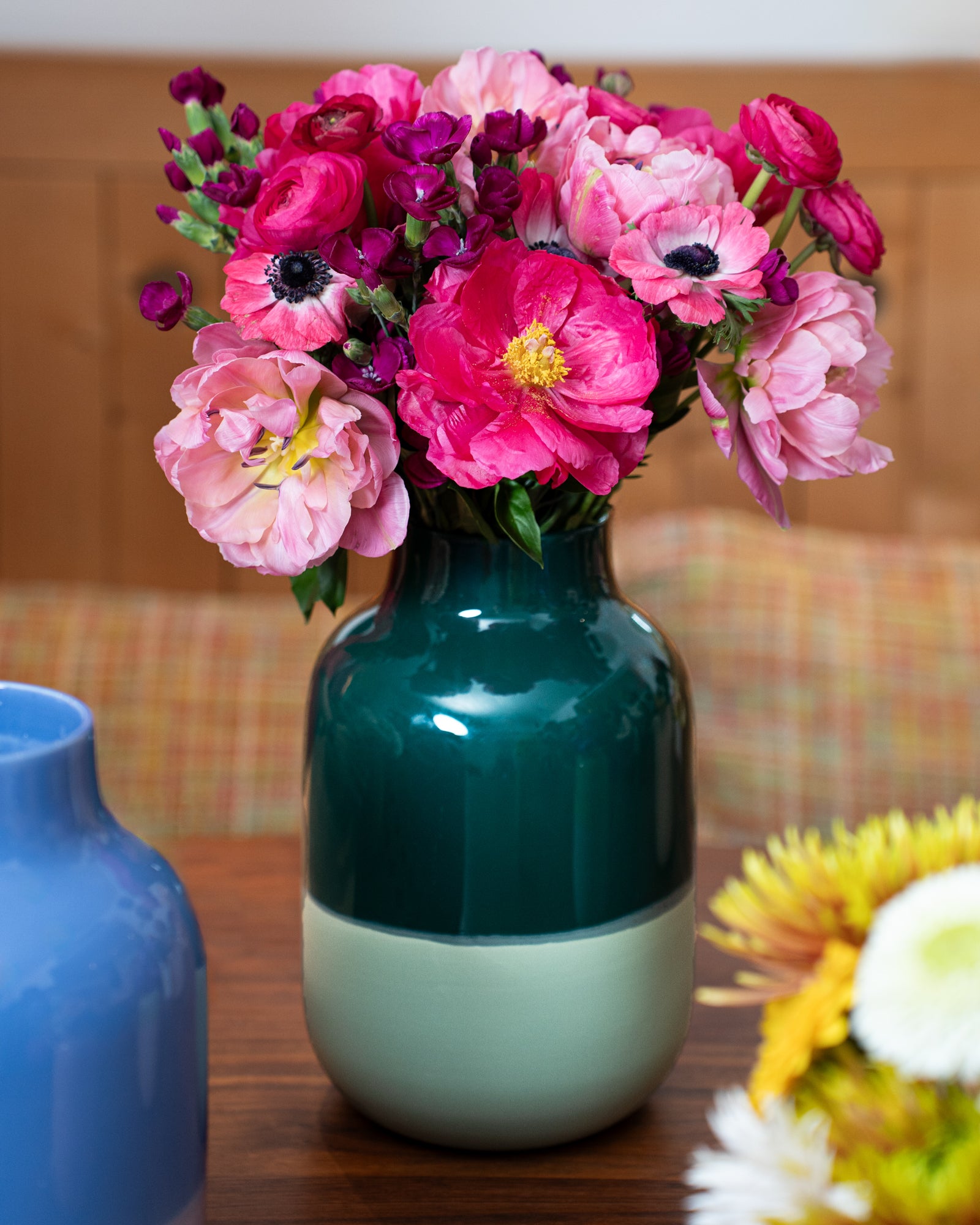 Ein Blumenstrauß mit rosa und roten Blüten in einer zweifarbigen Vase steht auf einem Tisch neben weiteren Vasen.