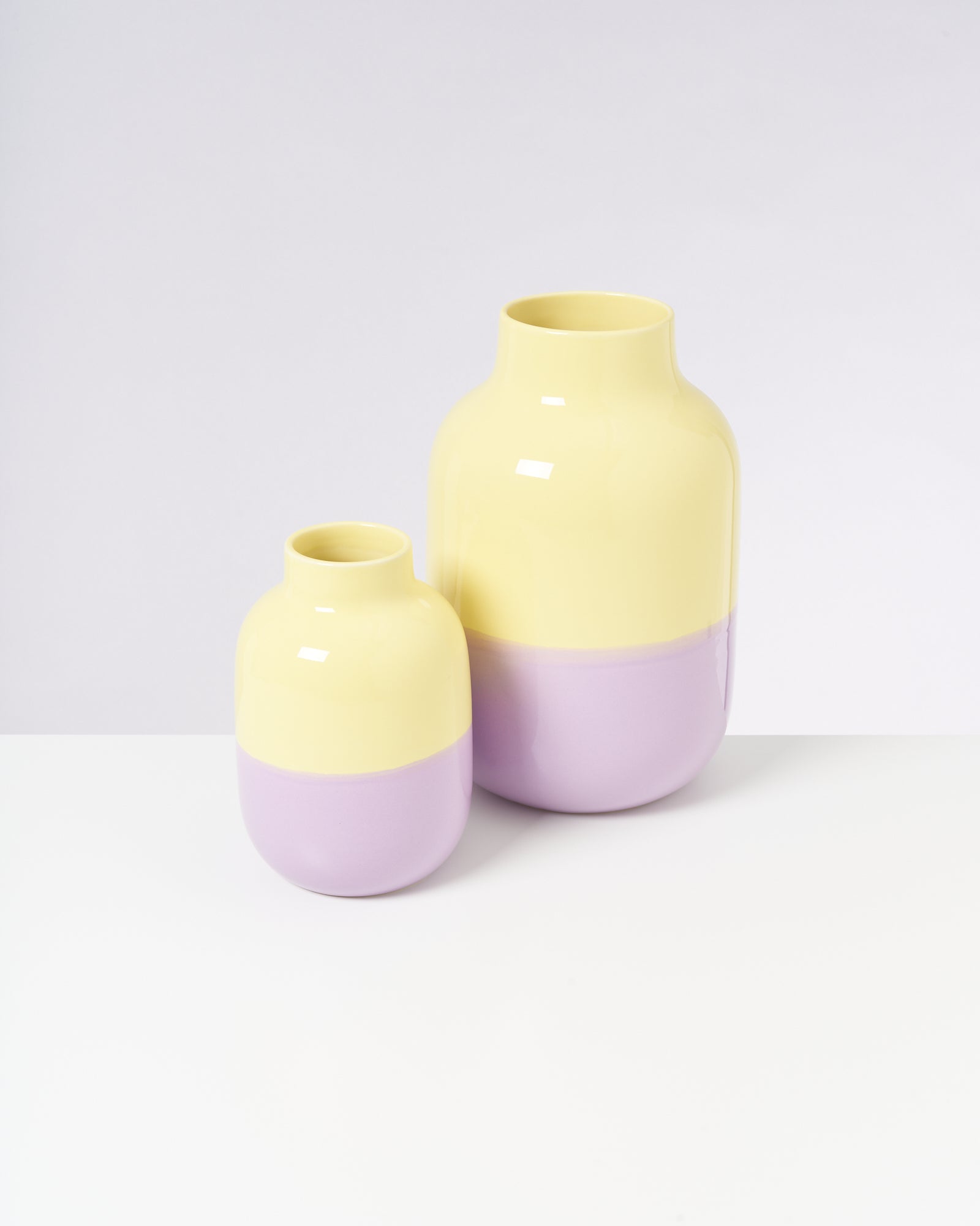 Zwei Vasen in Gelb und Lila stehen auf einer weißen Fläche vor einem hellgrauen Hintergrund.