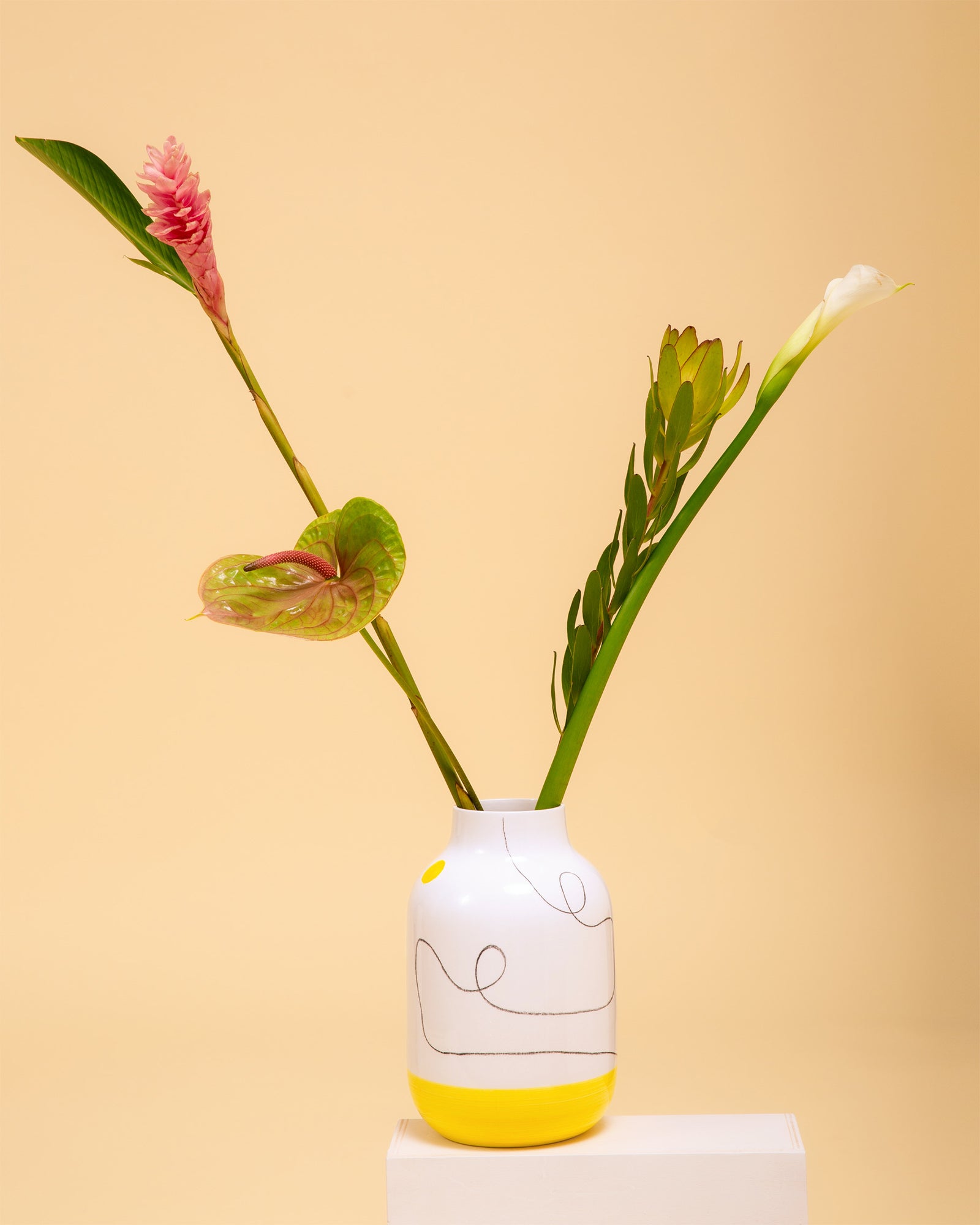 Weiße Vase mit gelbem Rand und abstraktem Muster, enthält rosafarbene und weiße Blumen, vor beigem Hintergrund.