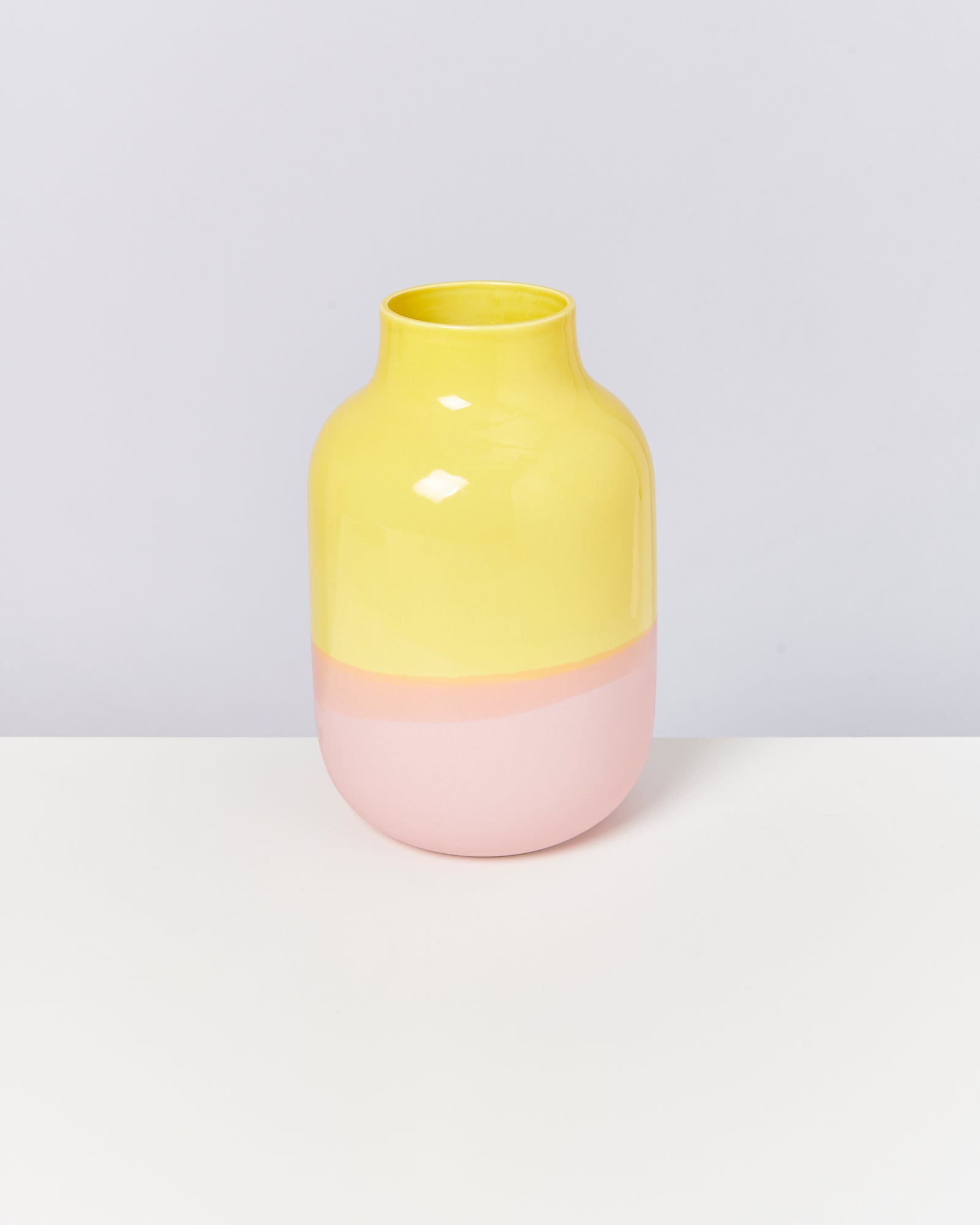 Zweifarbige Vase mit gelbem oberen und rosa unteren Teil steht auf einem weißen Tisch vor grauem Hintergrund.