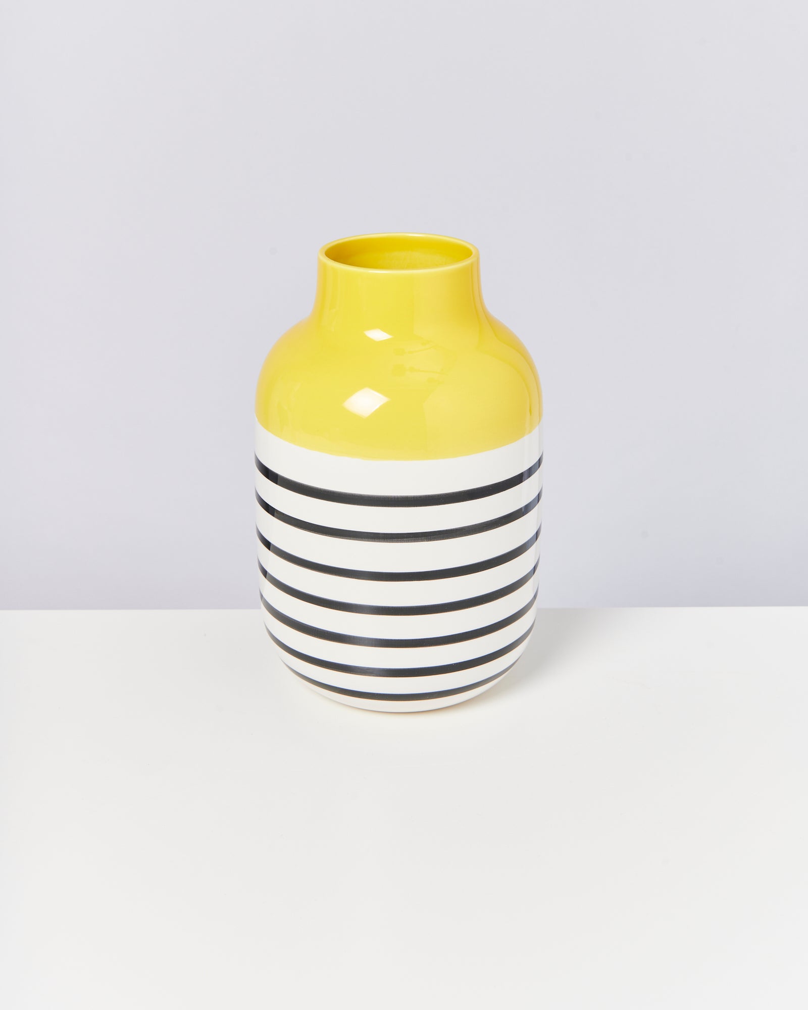 Gelbe Vase mit schwarzen und weißen Streifen steht auf einem weißen Untergrund vor grauem Hintergrund.