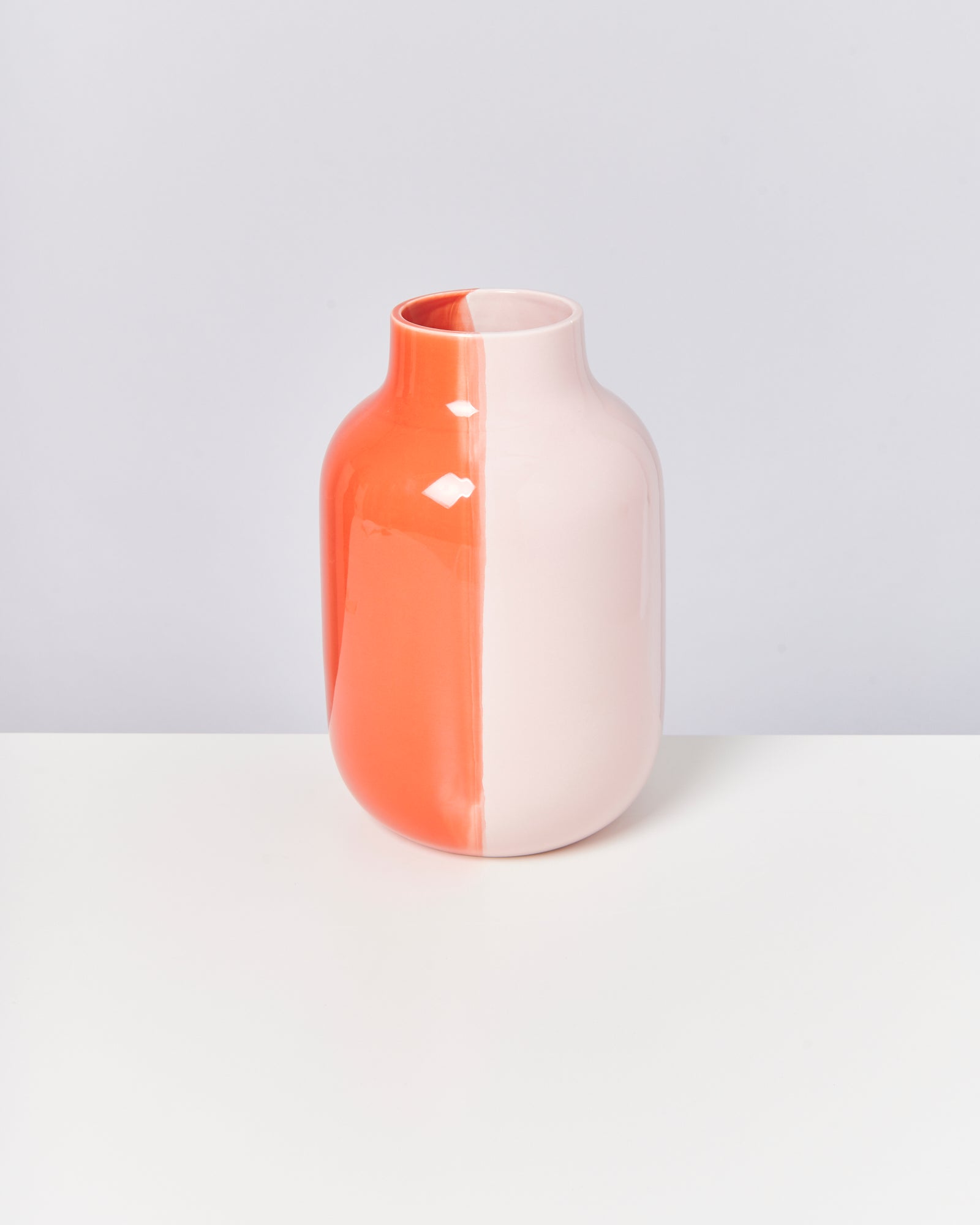 Zweifarbige Vase, halb orange, halb weiß, auf einem weißen Tisch vor grauem Hintergrund.