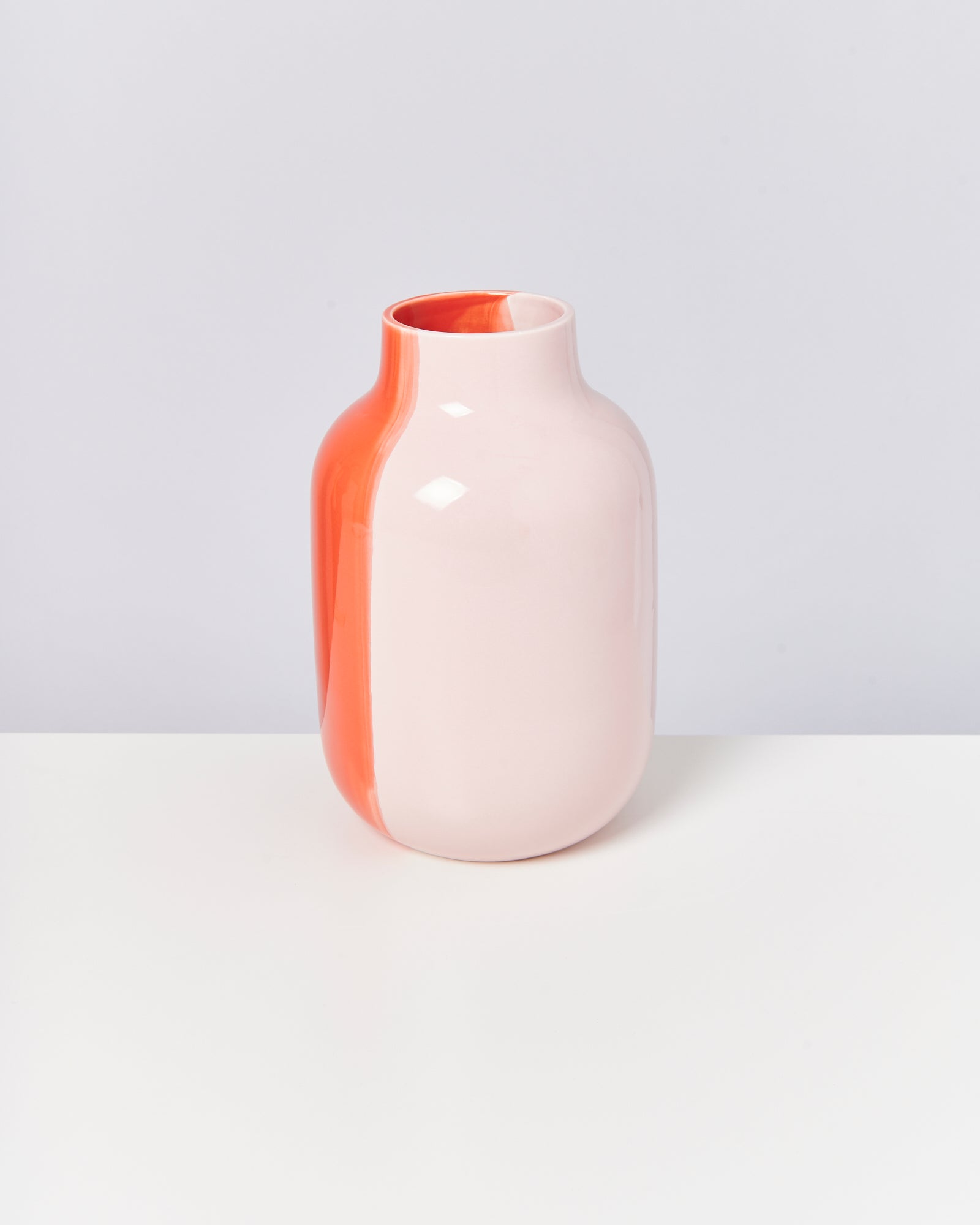 Eine zweifarbige Vase in Weiß und Orange steht auf einem weißen Tisch vor einem grauen Hintergrund.