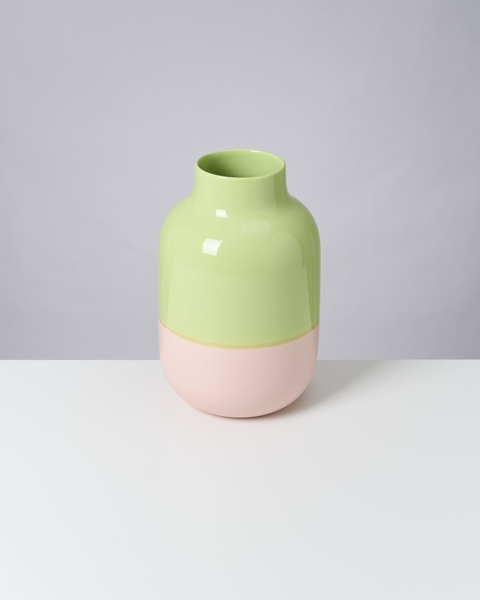 Zweifarbige Vase in Hellgrün und Rosa steht auf einem weißen Tisch vor grauem Hintergrund.