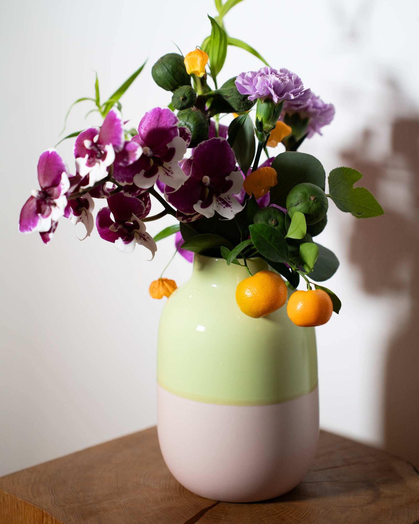 Bunter Blumenstrauß mit Orchideen und kleinen Orangen in einer zweifarbigen Vase auf einem Holztisch.