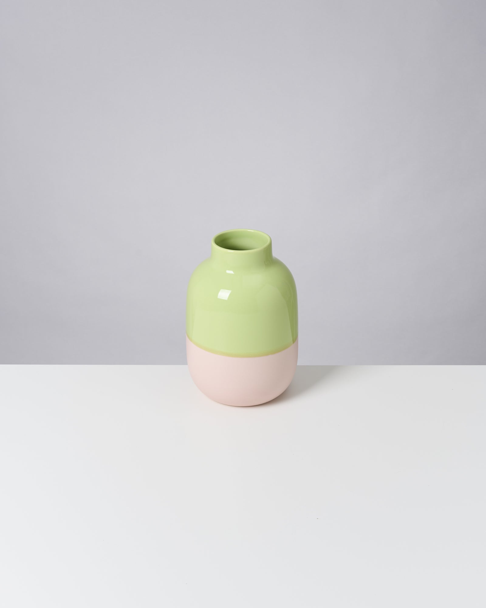 Zweifarbige Vase in Hellgrün und Rosa auf weißem Tisch vor grauem Hintergrund.
