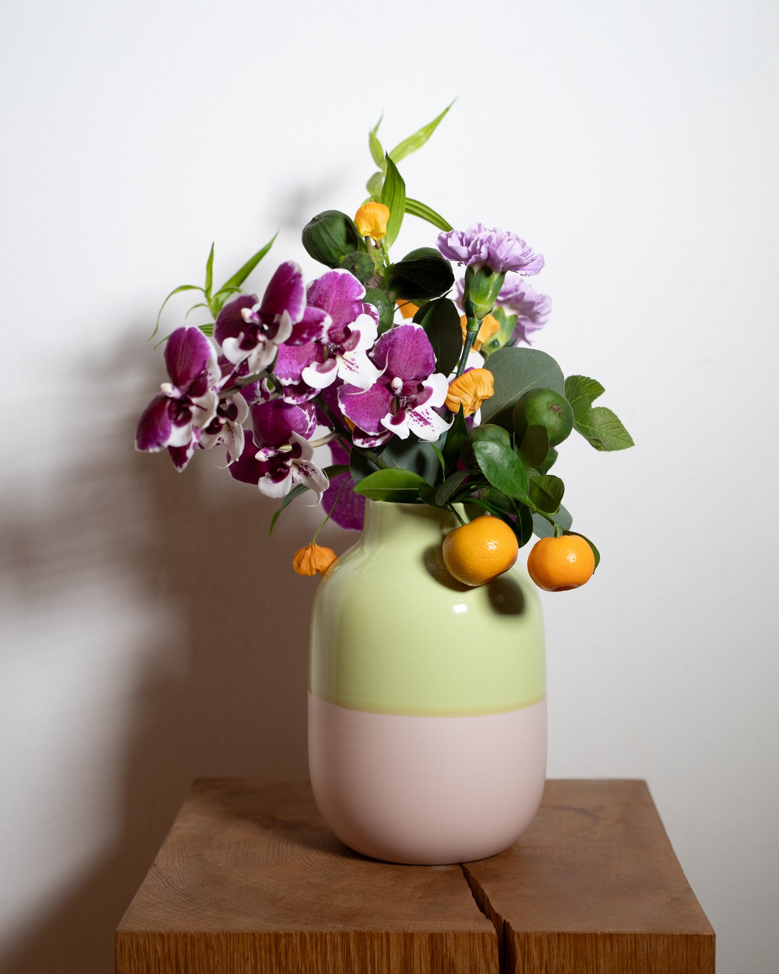 Bunter Blumenstrauß mit Orchideen und kleinen Früchten in einer zweifarbigen Vase auf einem Holzbrett.
