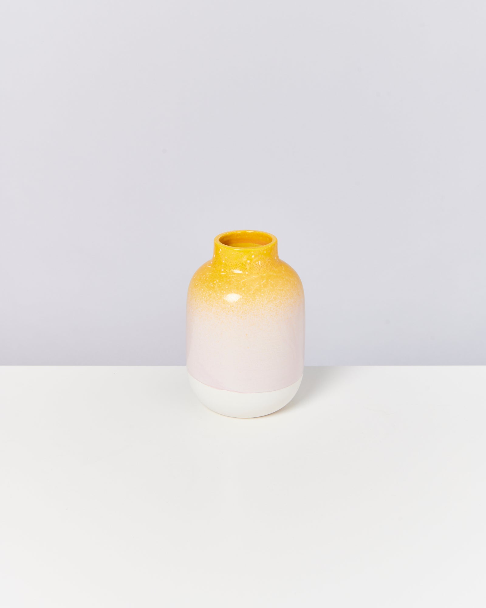 Gelb-weiß verlaufende Keramikvase mit glatter Oberfläche steht auf einem weißen Tisch vor grauem Hintergrund.