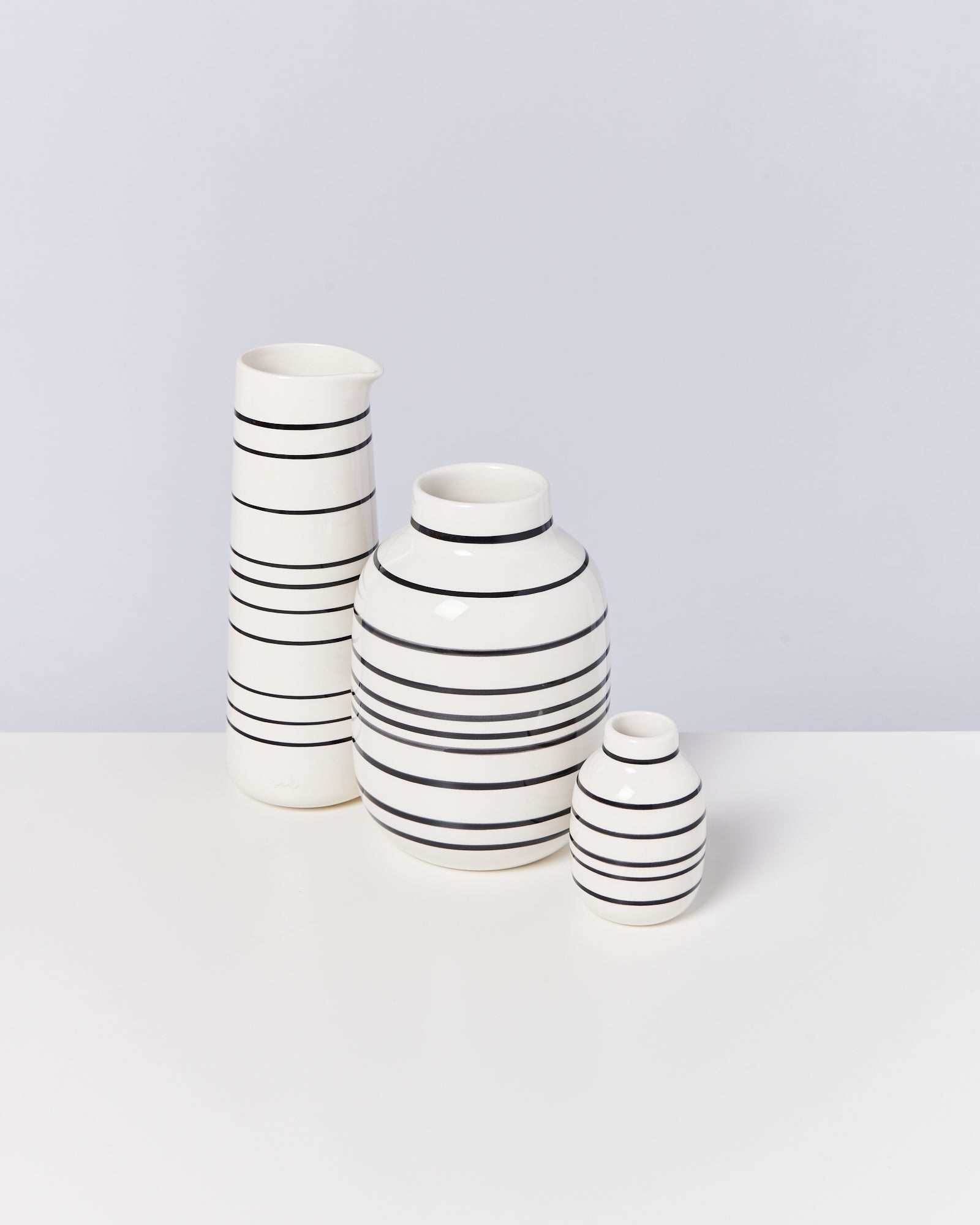 Drei weiß-schwarze, gestreifte Keramikkrüge in unterschiedlichen Größen stehen auf einem weißen Tisch.
