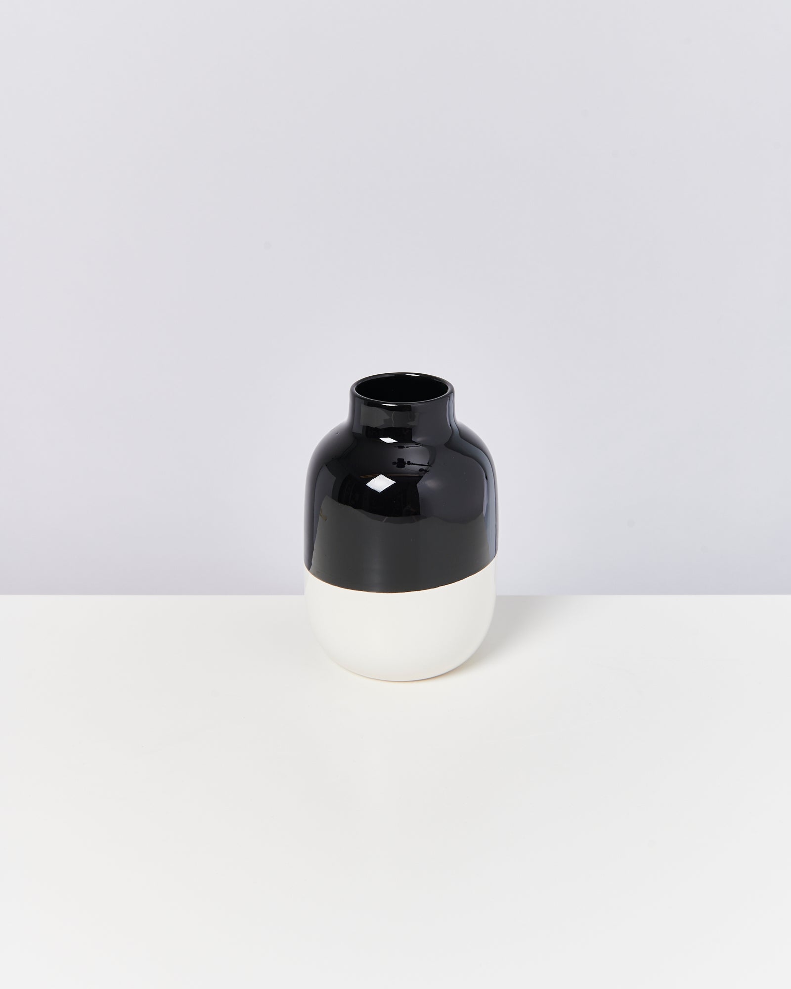 Eine bauchige Vase mit glänzend schwarzem oberen Teil und mattem weißen unteren Teil steht auf einem weißen Tisch.
