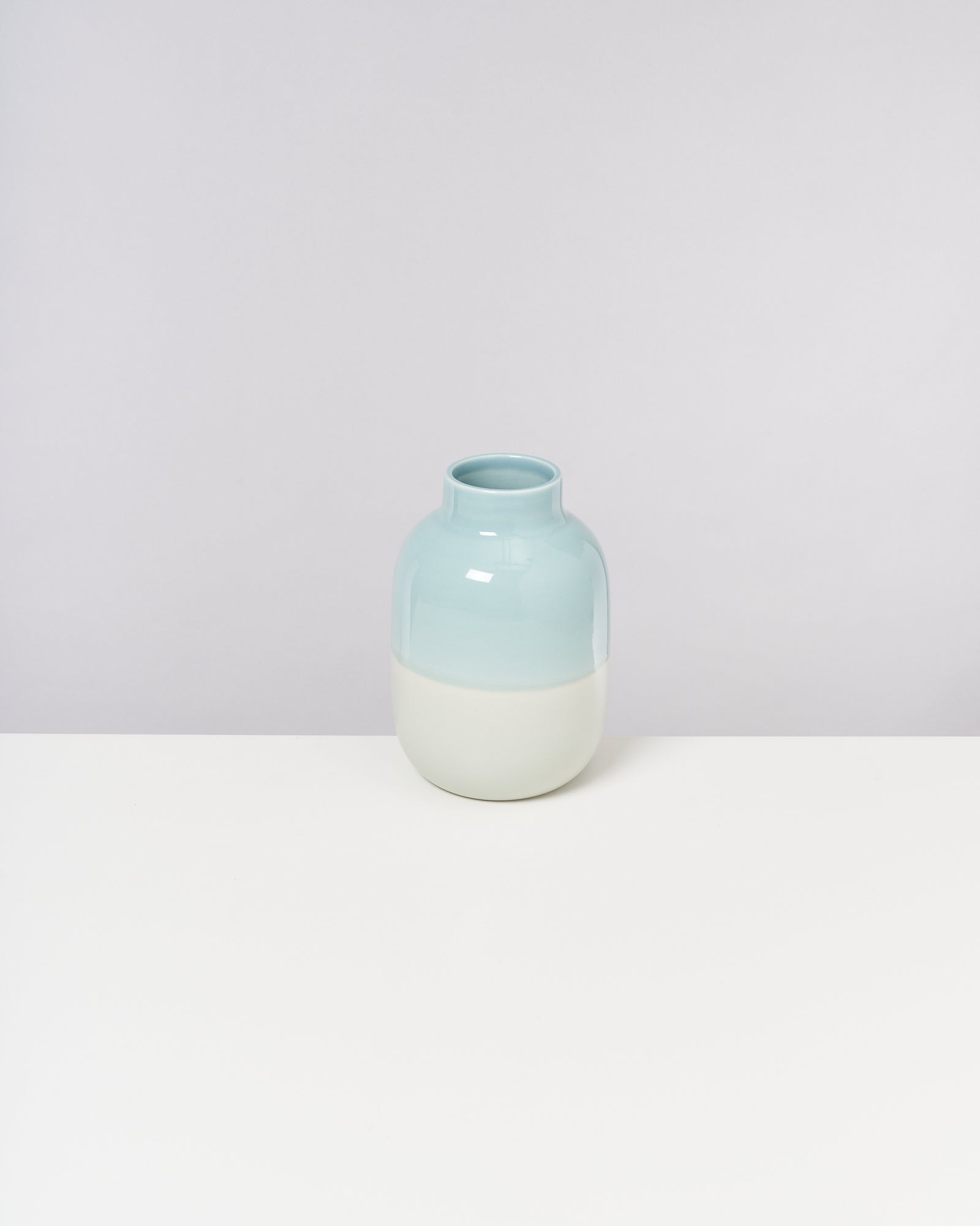Eine zweifarbige Vase in Hellblau und Weiß steht auf einem weißen Tisch vor einem grauen Hintergrund.
