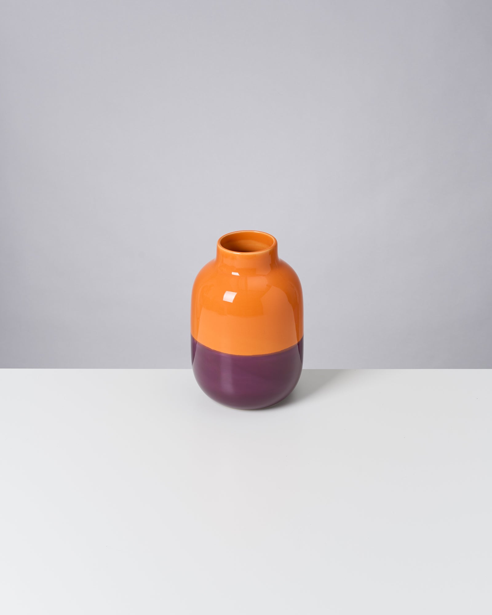 Orange- und lilafarbene Vase steht auf einem weißen Tisch vor einem grauen Hintergrund.