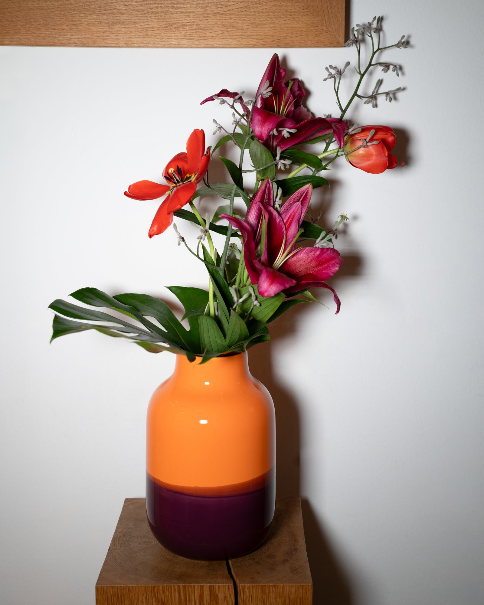 Eine orange-violette Vase mit roten und pinken Blumen steht auf einem Holztisch vor einer weißen Wand.