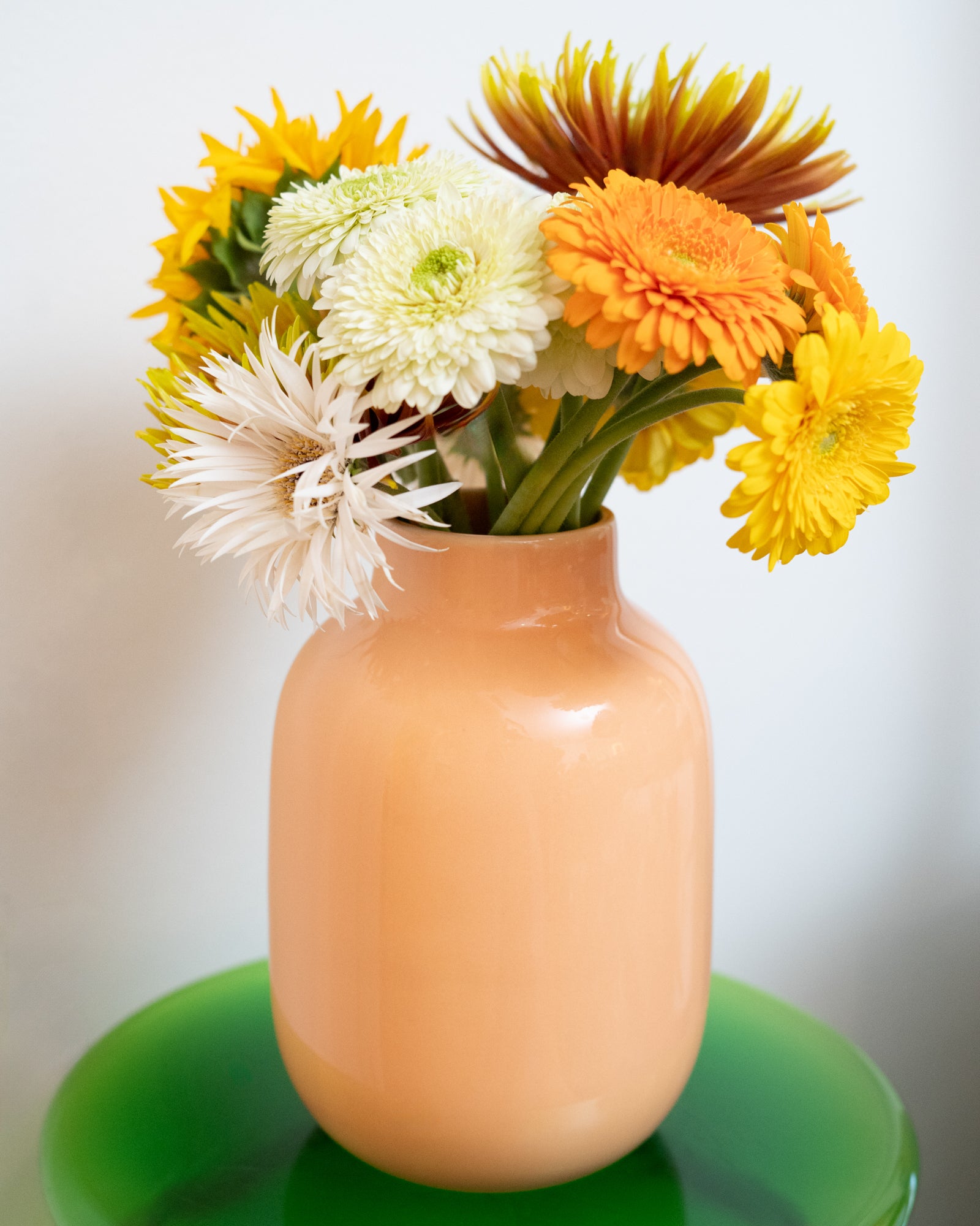 Ein blumiger Strauß mit gelben, weißen und orangefarbenen Blüten in einer pfirsichfarbenen Vase auf einem grünen Tisch.