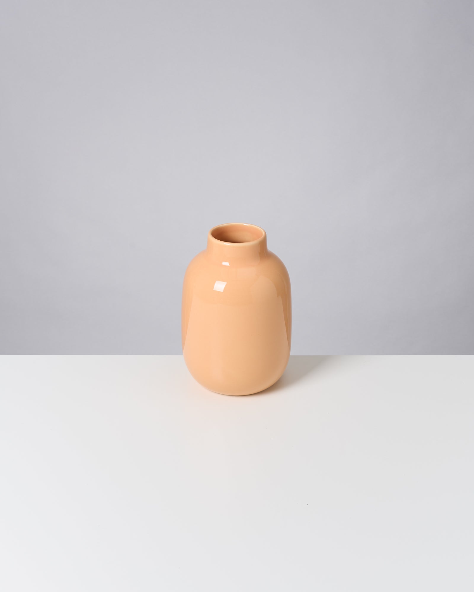 Beigefarbene Keramikvase steht auf einem weißen Tisch vor einem grauen Hintergrund.