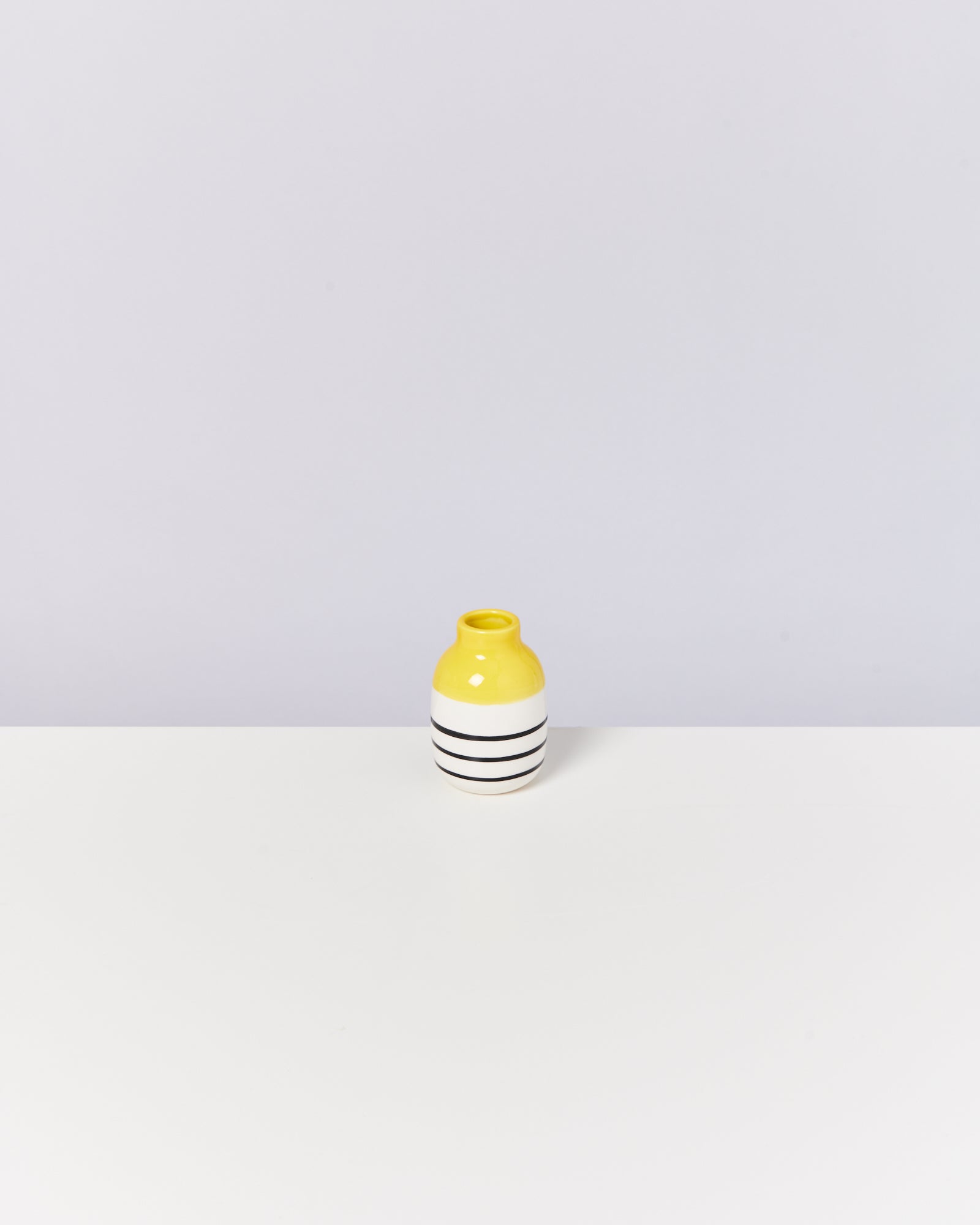 Gelbe Vase mit schwarzen Streifen auf weißer Oberfläche vor grauem Hintergrund.