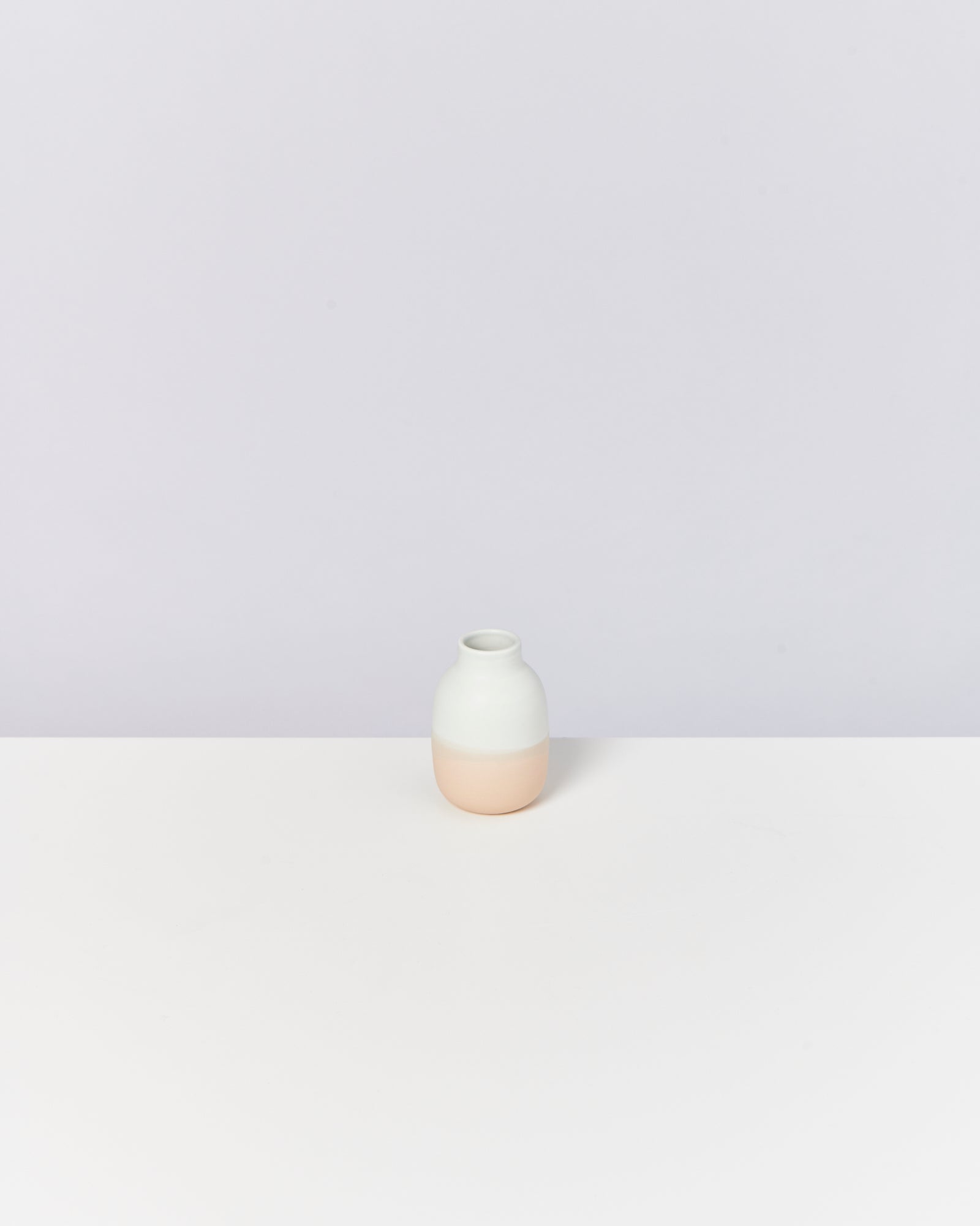 Kleine, zweifarbige Vase mit weißem oberen und beigem unteren Teil auf einer weißen Oberfläche vor grauem Hintergrund.
