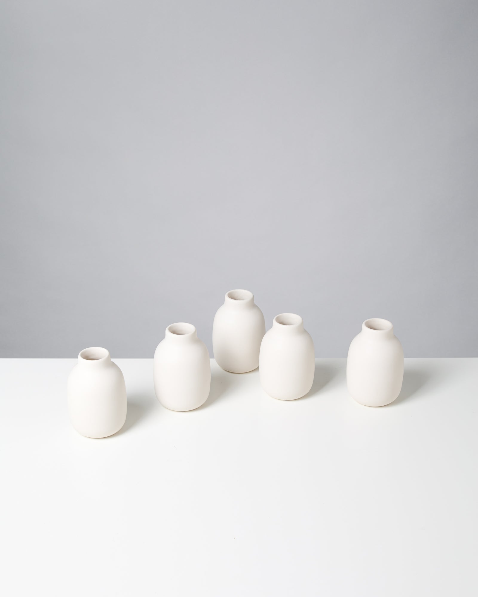 Fünf weiße, bauchige Keramikvasen stehen auf einem weißen Tisch vor einem grauen Hintergrund.