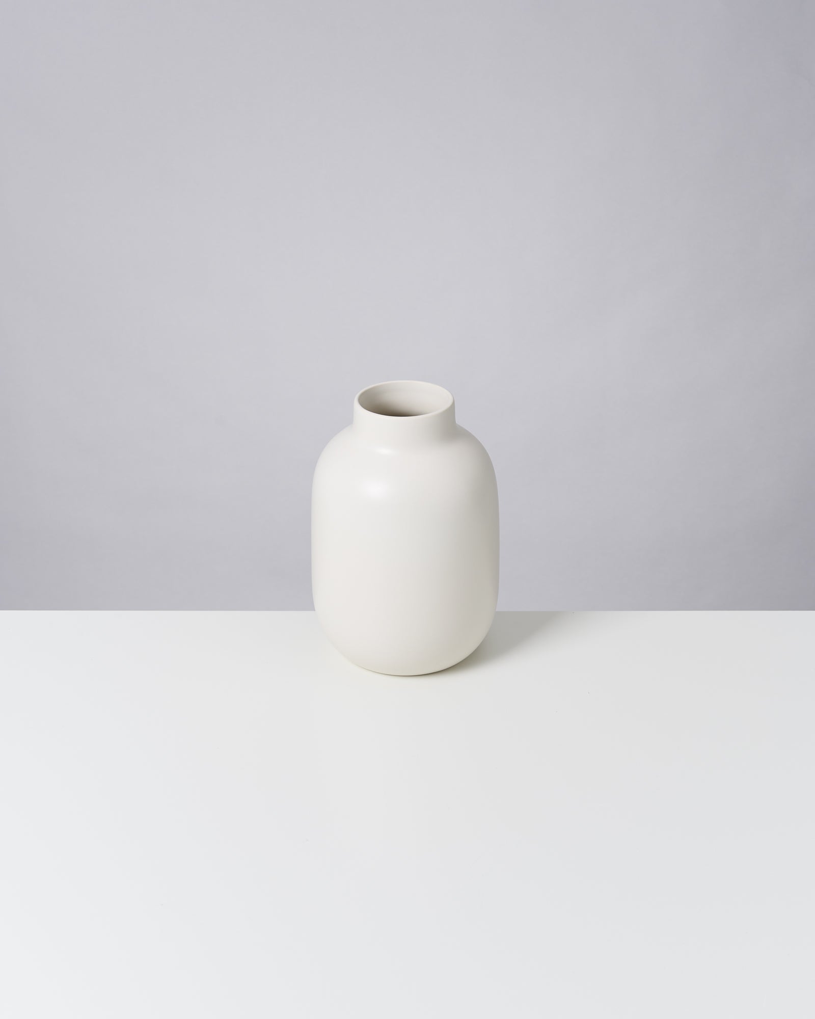 Weiße, minimalistische Vase auf einem weißen Tisch vor grauem Hintergrund.