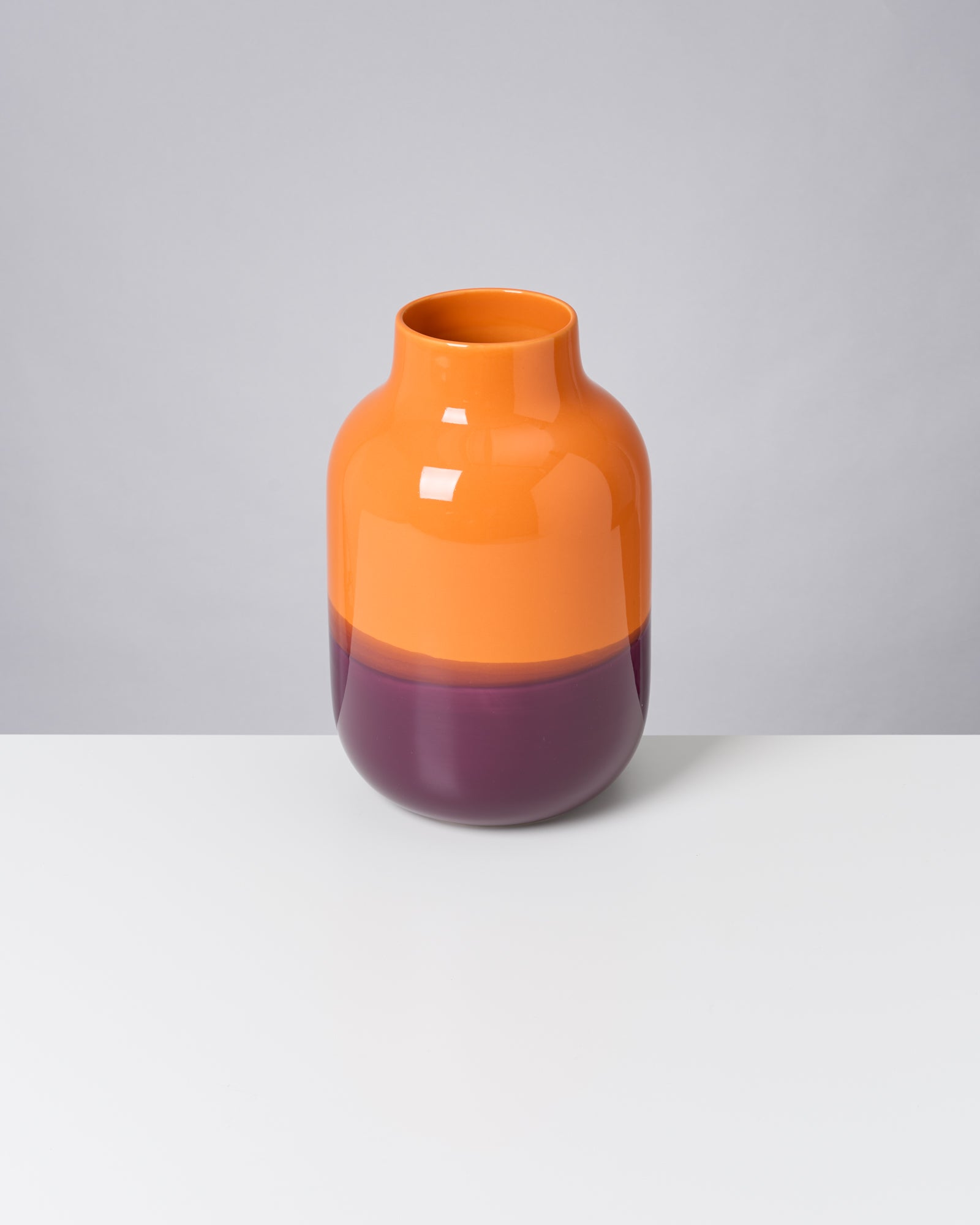 Zweifarbige Vase in Orange und Lila auf einem weißen Tisch vor grauem Hintergrund.