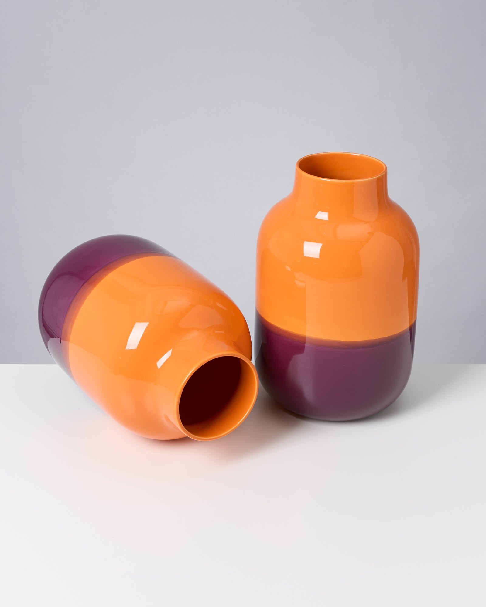 Zwei Vasen in Orange und Lila, eine stehend und eine liegend, auf weißem Hintergrund.