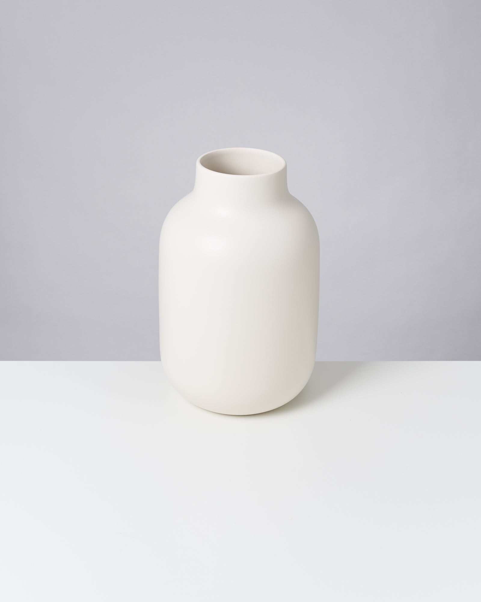Weiße, minimalistische Vase auf einem weißen Tisch vor grauem Hintergrund.