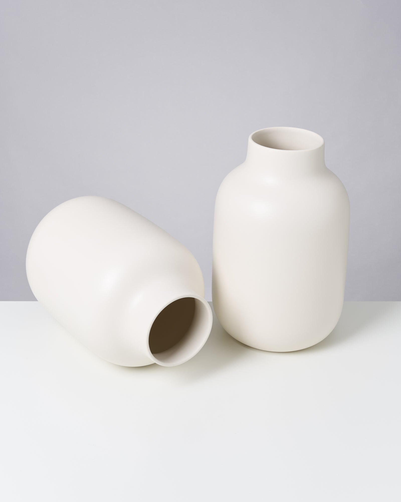 Zwei cremefarbene Vasen, eine aufrecht und eine liegend, auf einer weißen Oberfläche vor grauem Hintergrund.