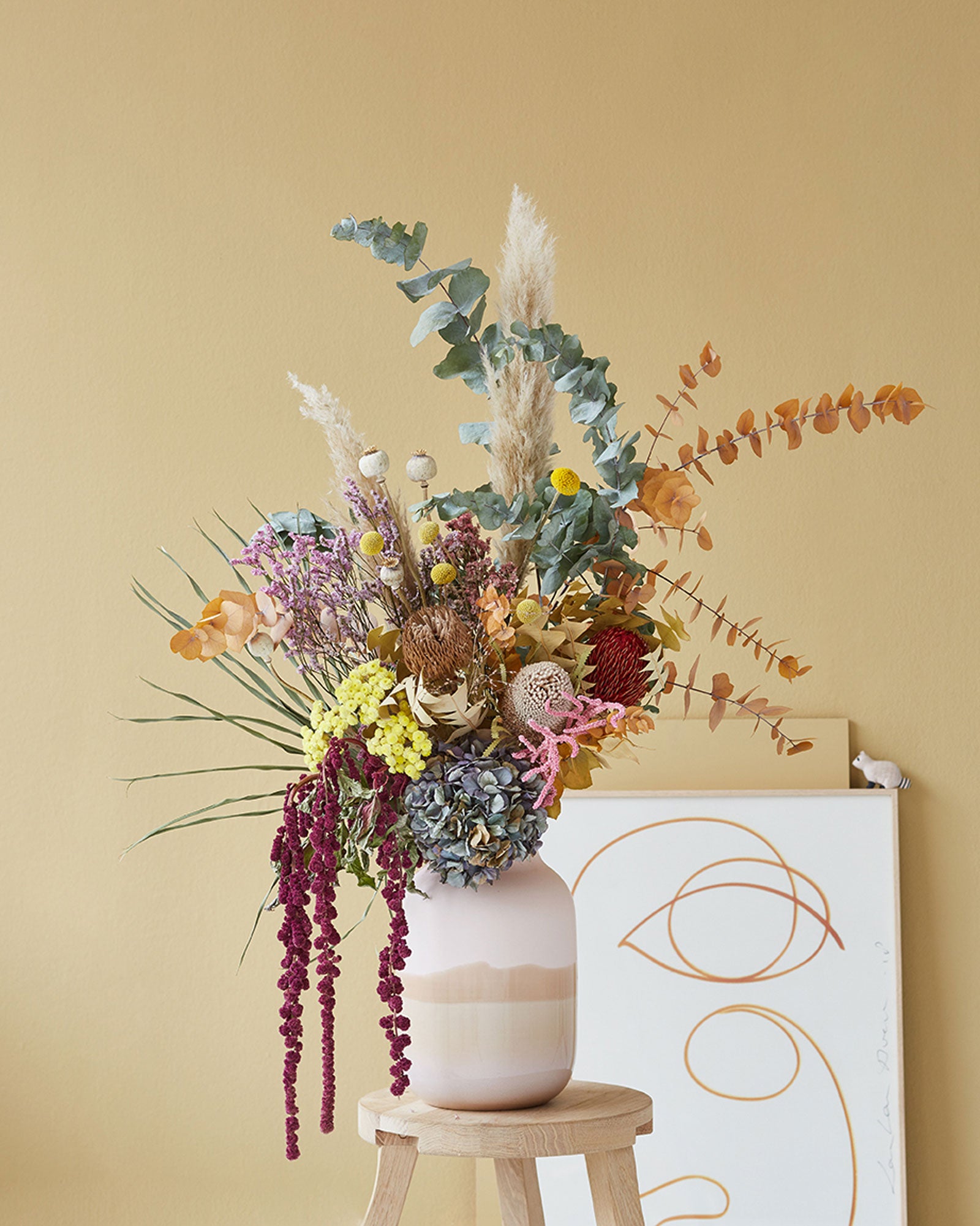 Ein bunter Strauß aus Trockenblumen in einer Vase auf einem Hocker vor einer beigen Wand und abstrakter Kunst.